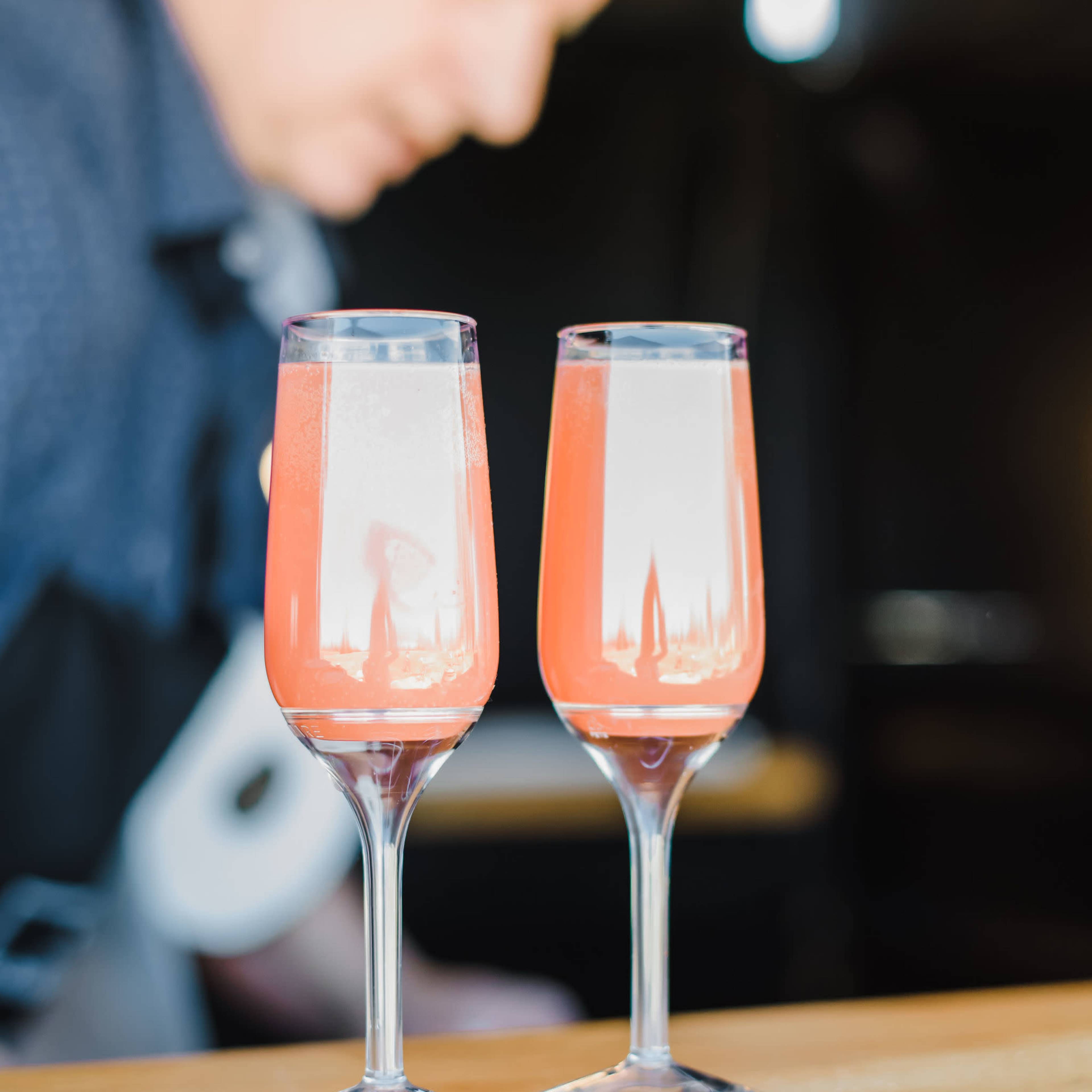 Two glasses of pink cocktails are placed on a wooden countertop, with a blurred figure preparing drinks in the background.