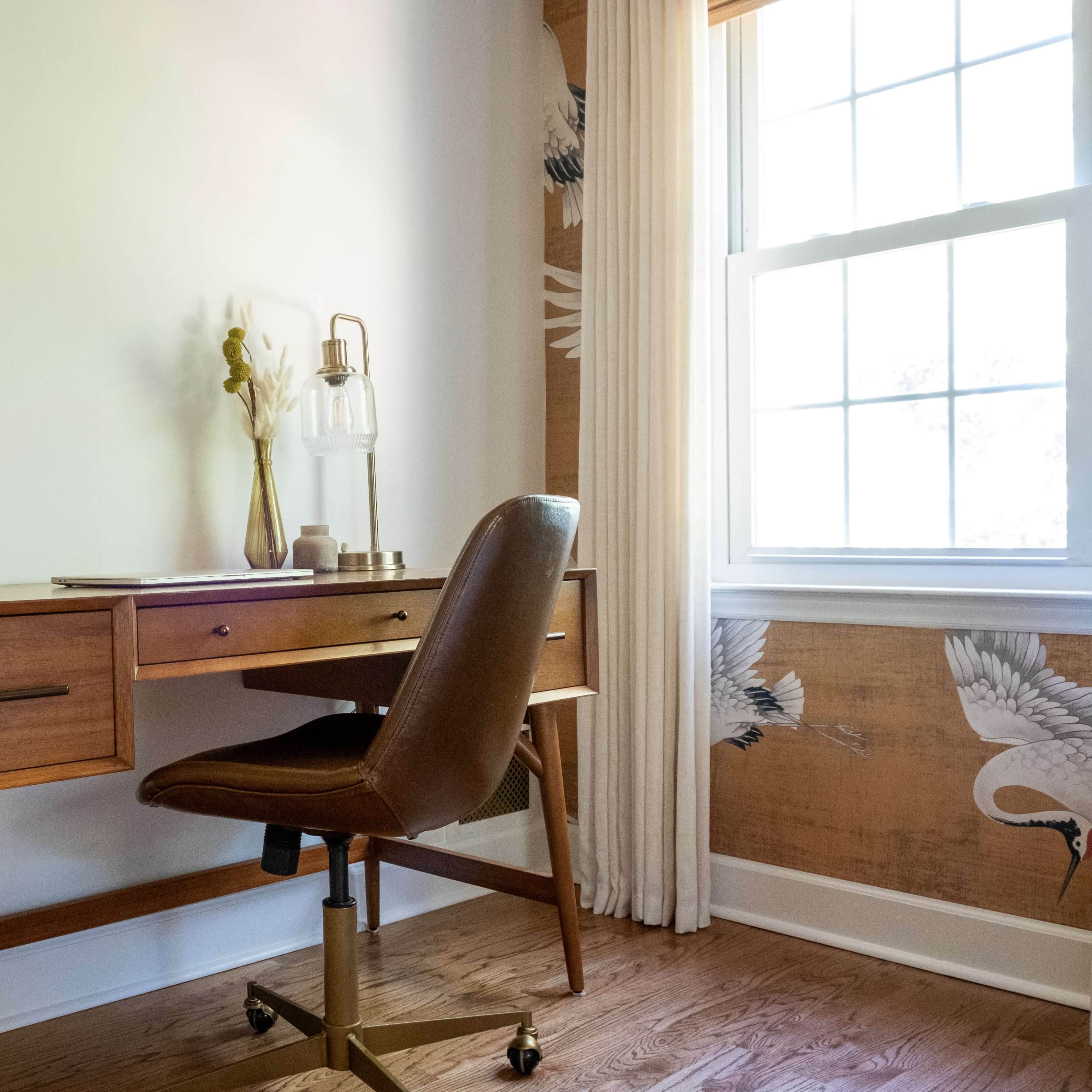 A wooden desk with a chair is positioned next to a window, featuring a lamp and decorative items, against a wall adorned with a nature-themed wallpaper.