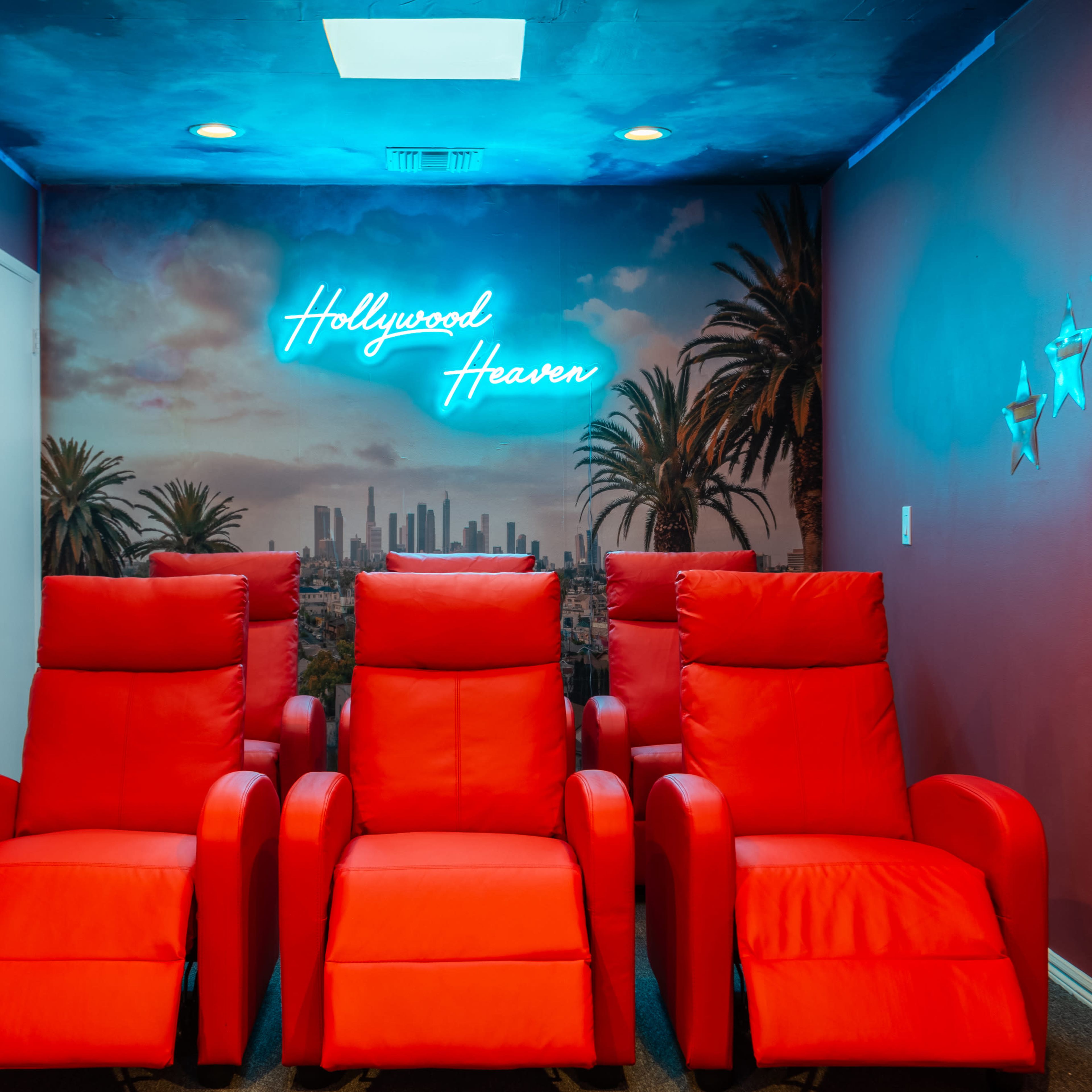A cozy home theater with four red reclining chairs in front of a wall featuring a neon sign that reads "Hollywood Heaven" and a mural of a city skyline framed by palm trees.