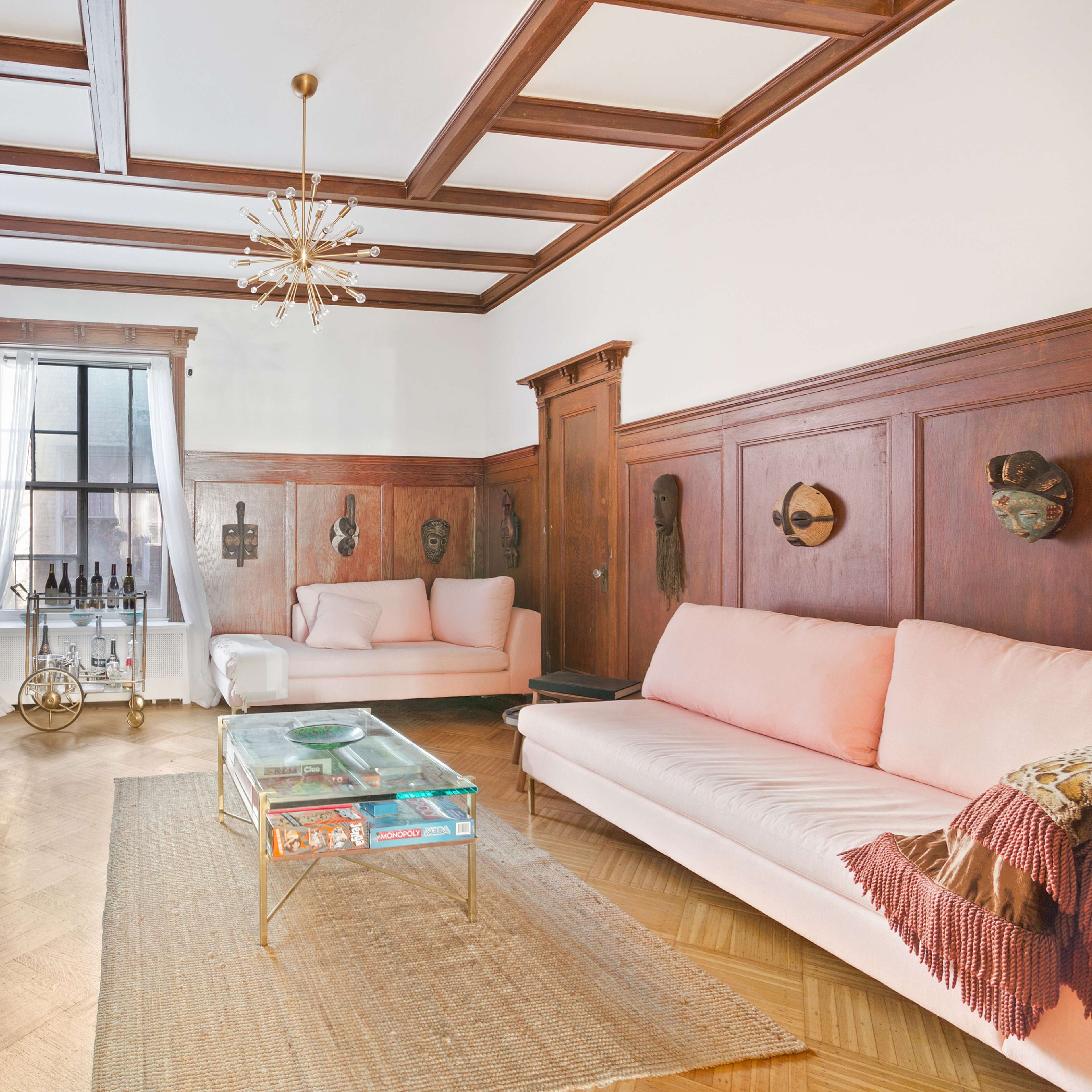 A living room with wood-paneled walls, a coffered ceiling, two pink sofas, a glass coffee table, and built-in bookshelves.