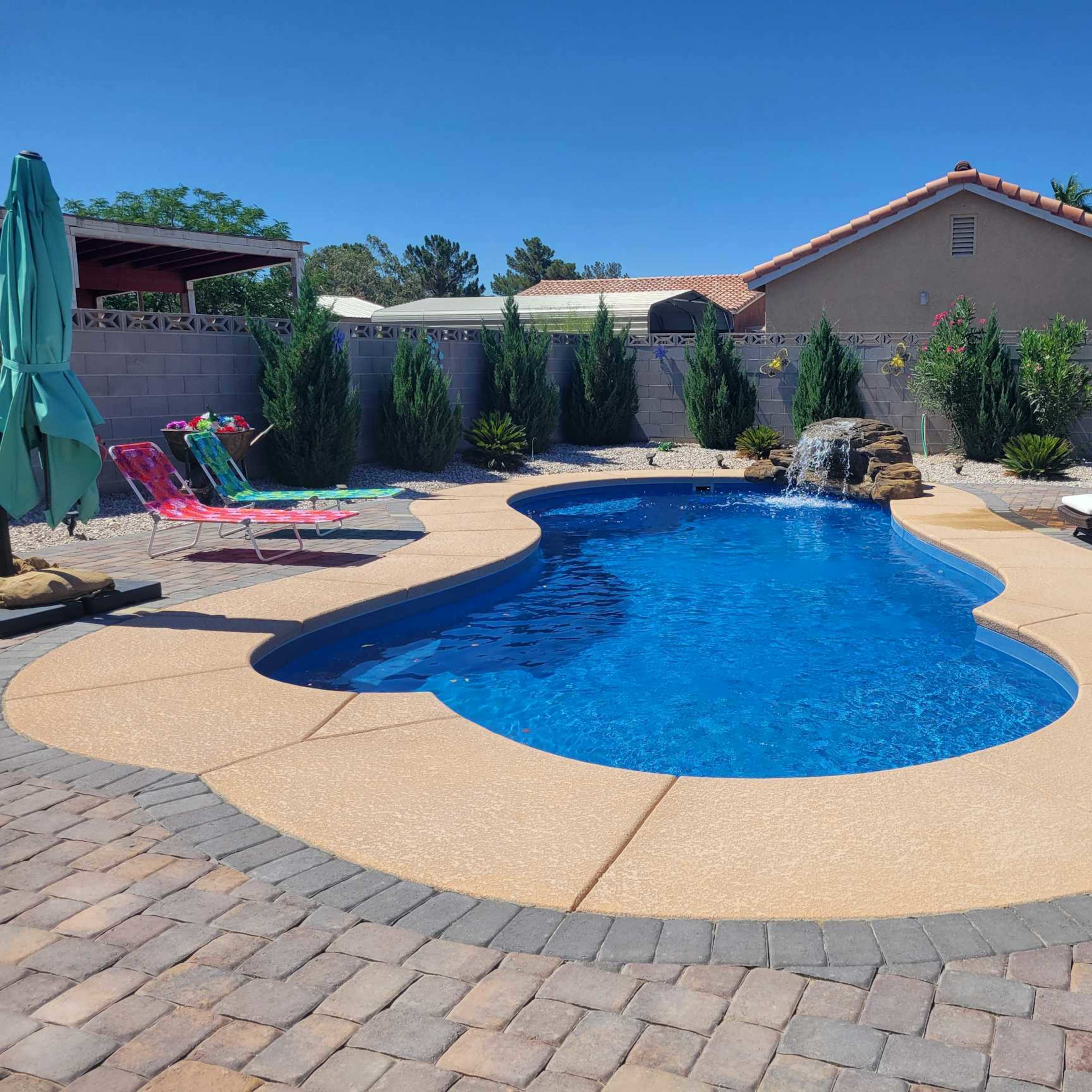A backyard features a curvy blue swimming pool surrounded by lounge chairs, umbrellas, and landscaped greenery.