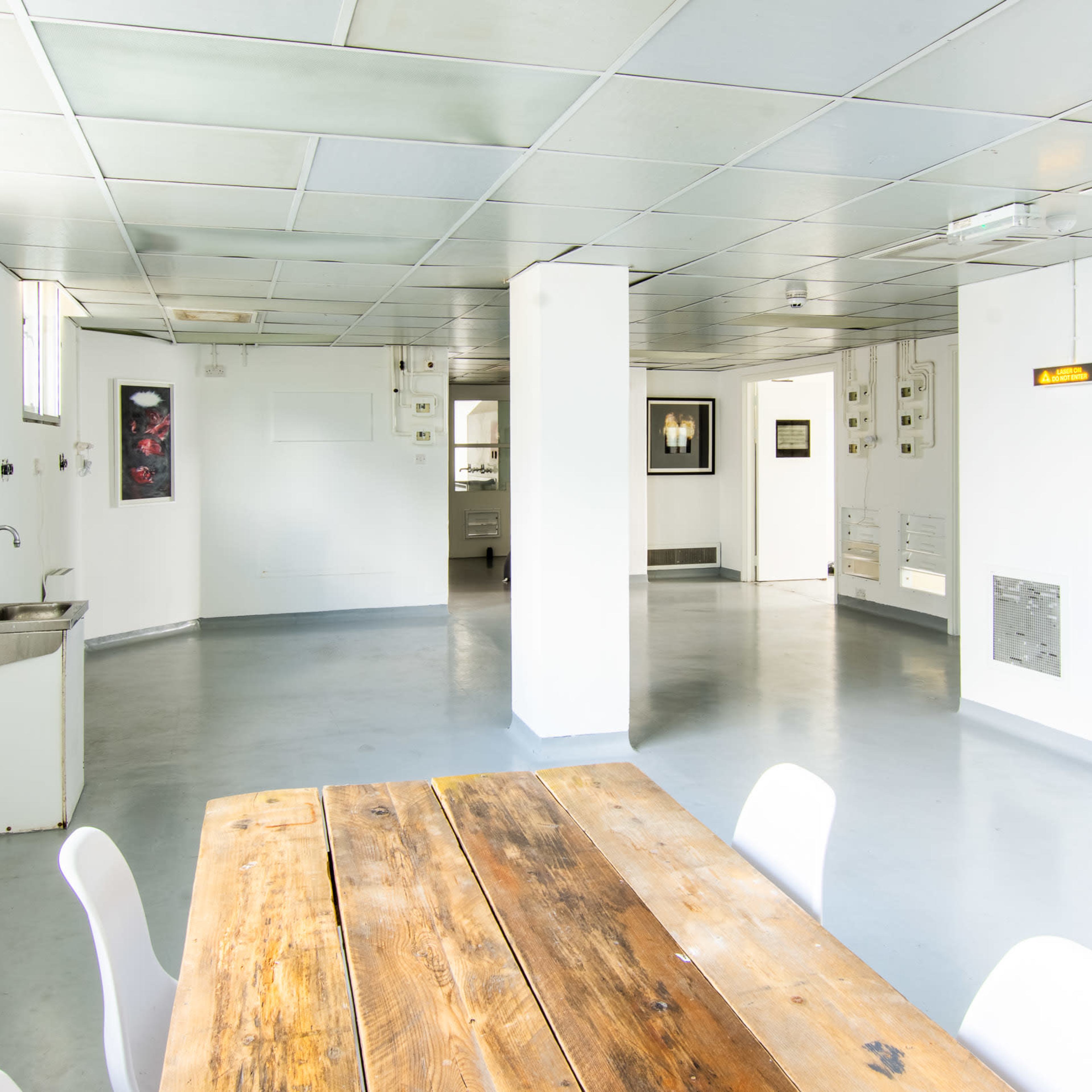 A spacious, well-lit room with a wooden table and white chairs, featuring kitchen facilities and multiple artwork displays on the walls.