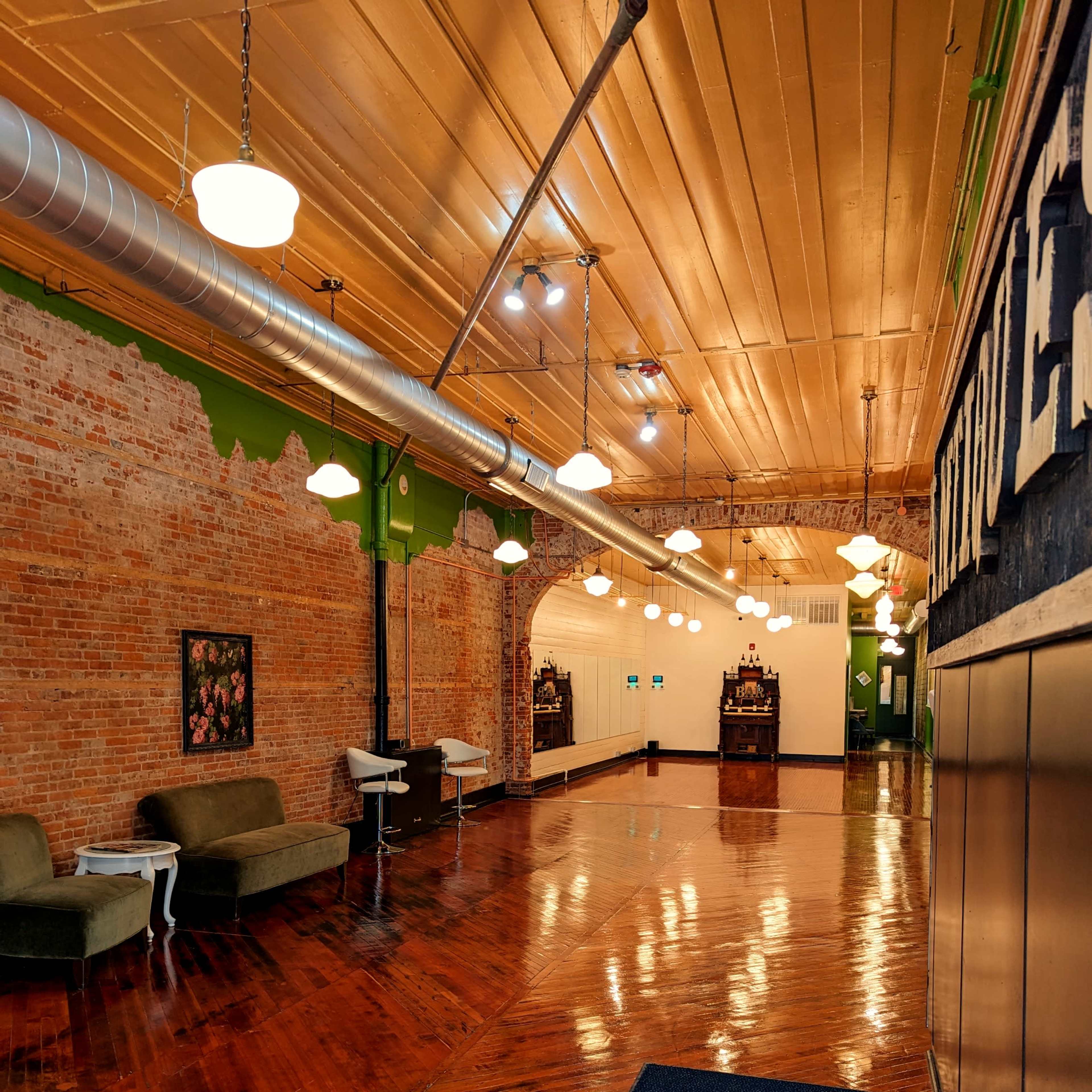 A wide hallway featuring exposed brick walls, polished wooden floors, and green accents, with seating and fixtures along the sides.