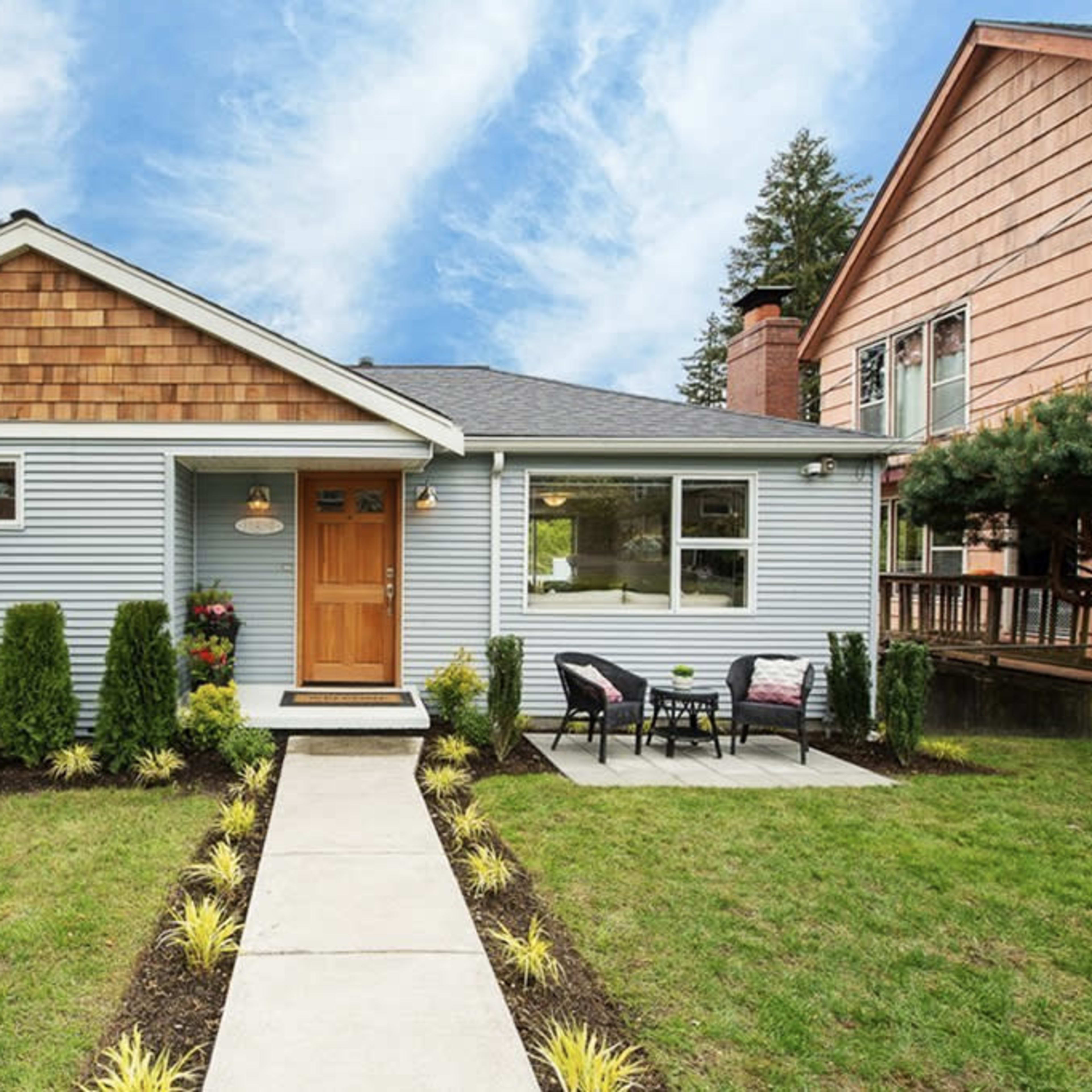 A single-story house with a light blue exterior, a wooden front door, and a neatly landscaped yard featuring a pathway and manicured plants.