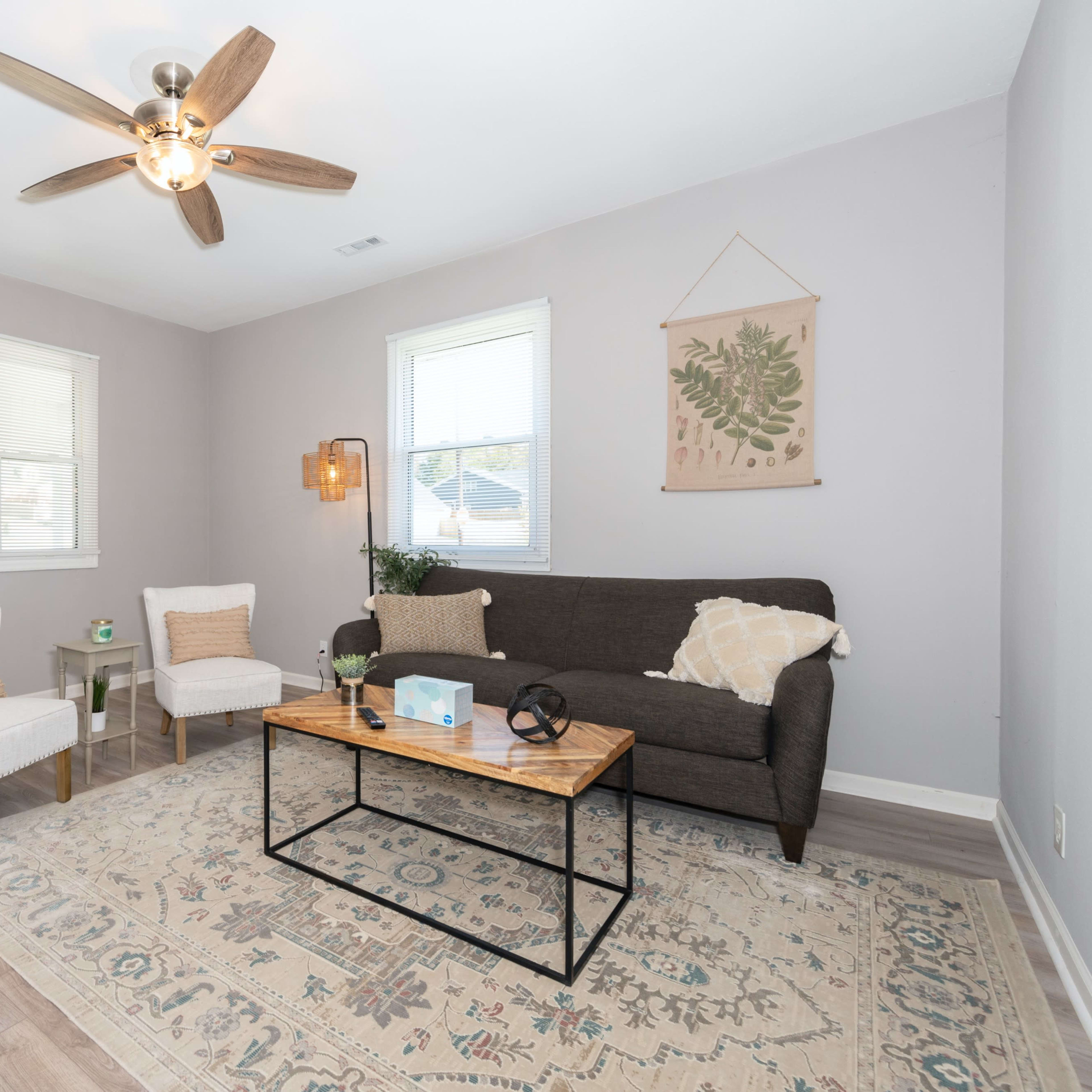 The living room features a brown sofa, two white chairs, a coffee table, and a ceiling fan, all arranged on a wooden floor with a decorative area rug.