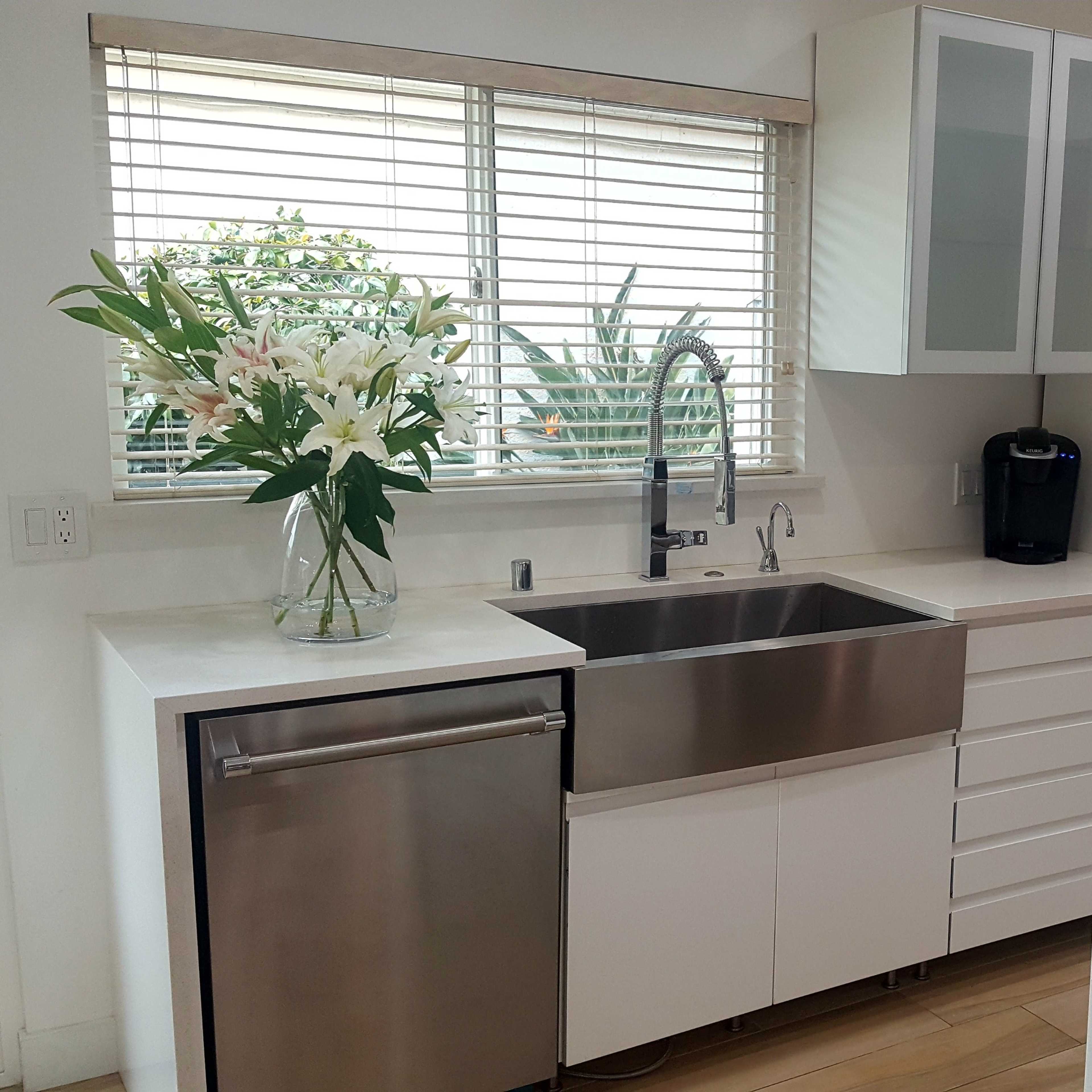 A modern kitchen with a stainless steel sink, white cabinetry, and a large window featuring floral arrangements.