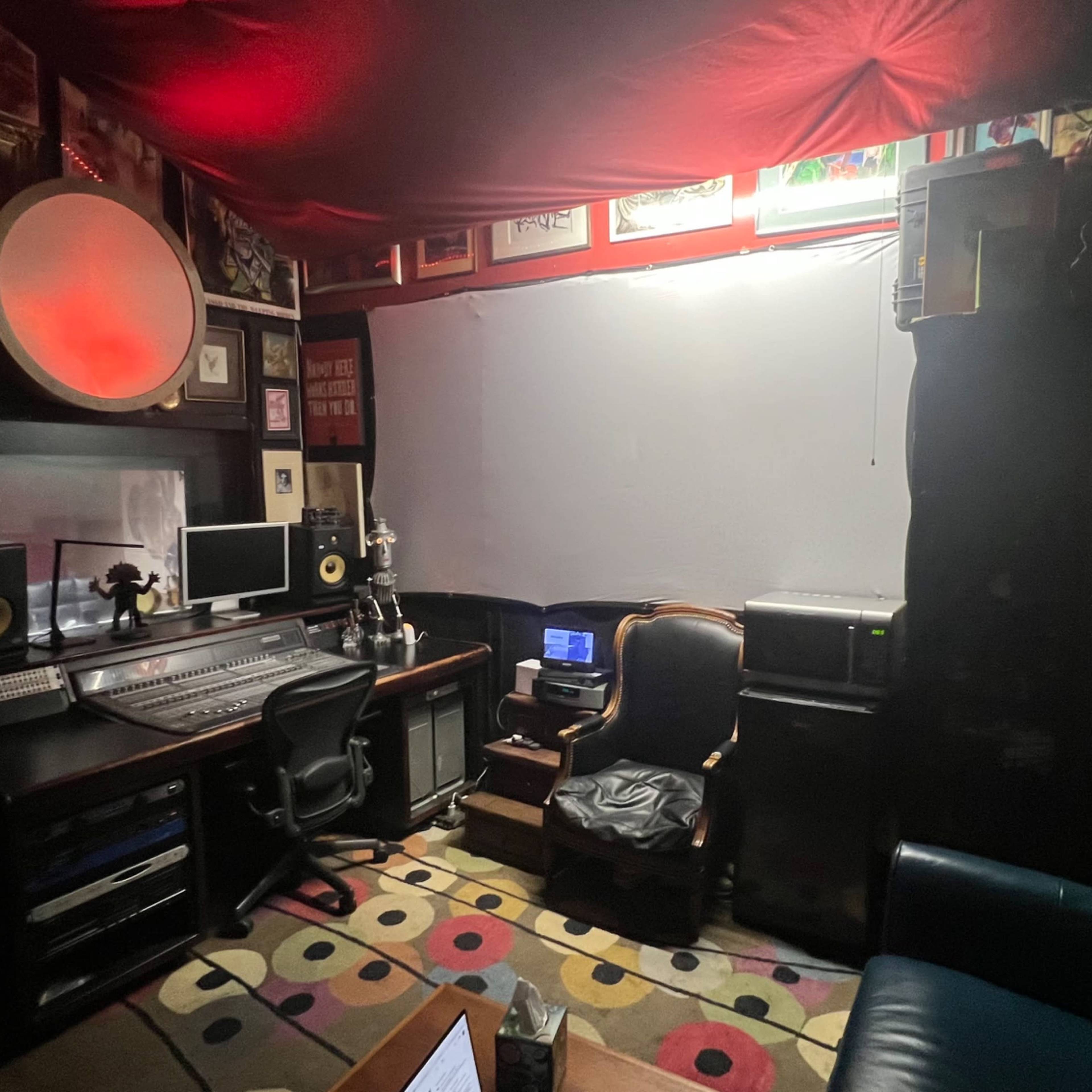 A dimly lit music studio with a mixing console, computer setup, an armchair, and various framed artworks on the walls.