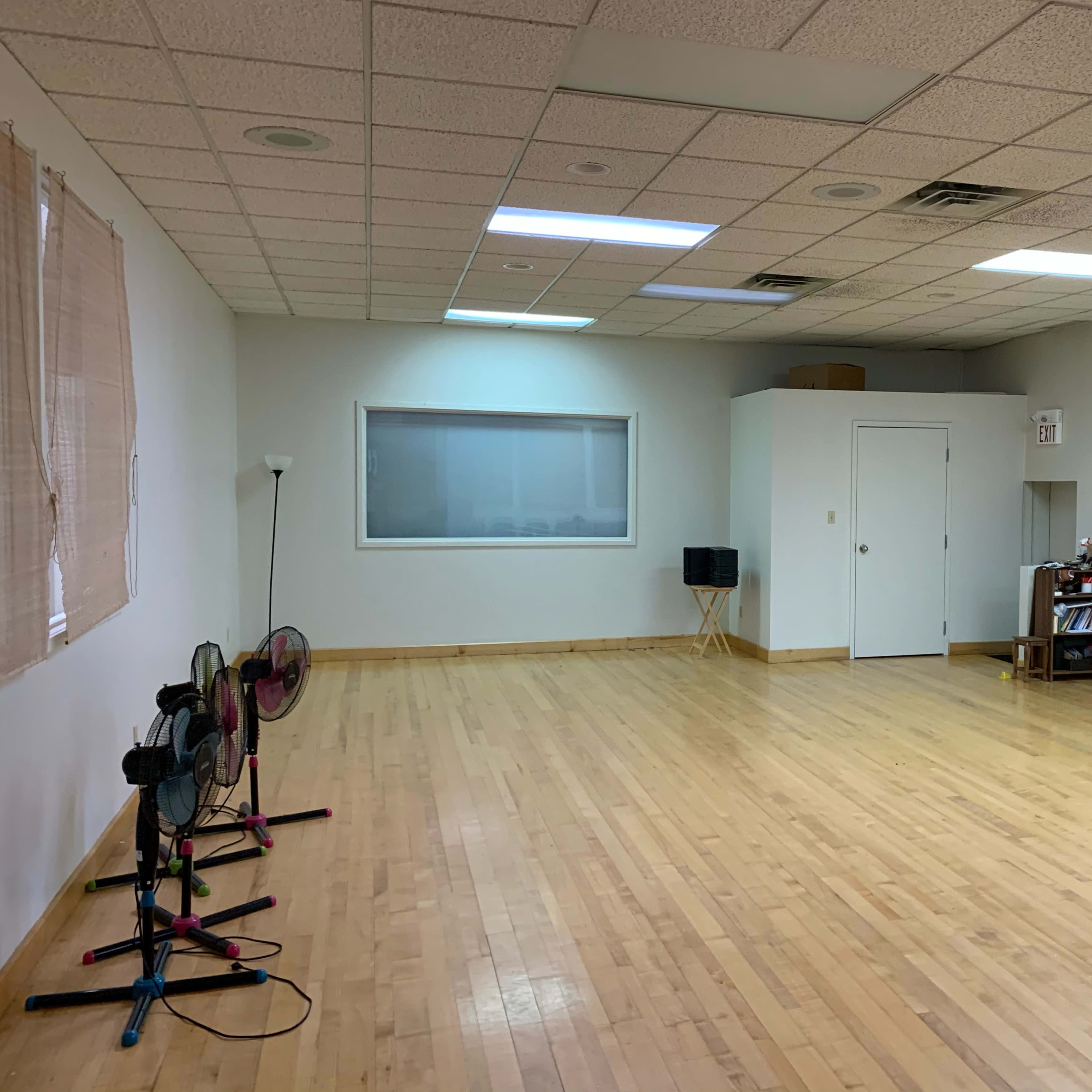 An empty room with wooden floors, a large window, and two standing fans positioned against one wall.