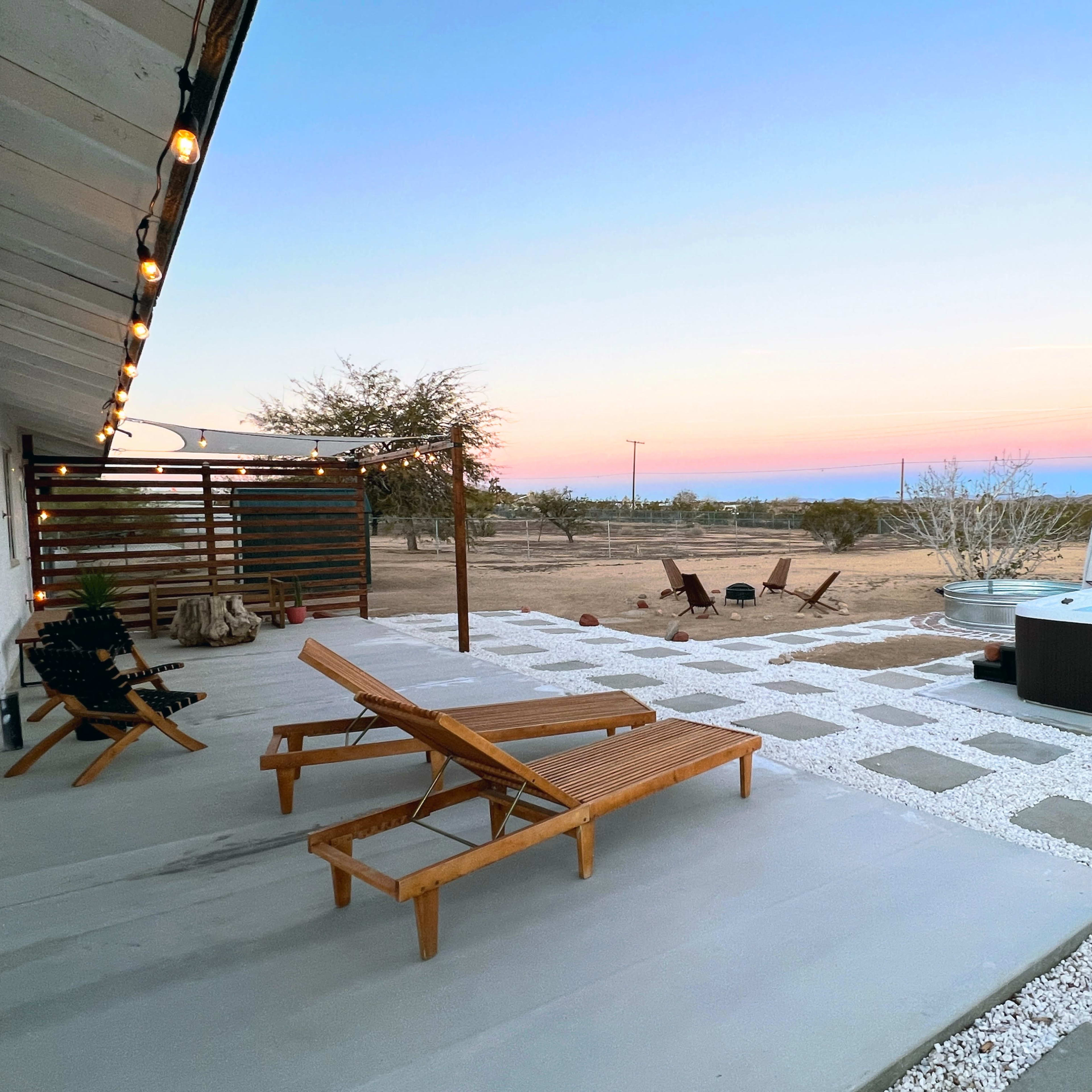 The scene shows a patio area with wooden lounge chairs, a hot tub, and a desert landscape under a pastel sunset sky.