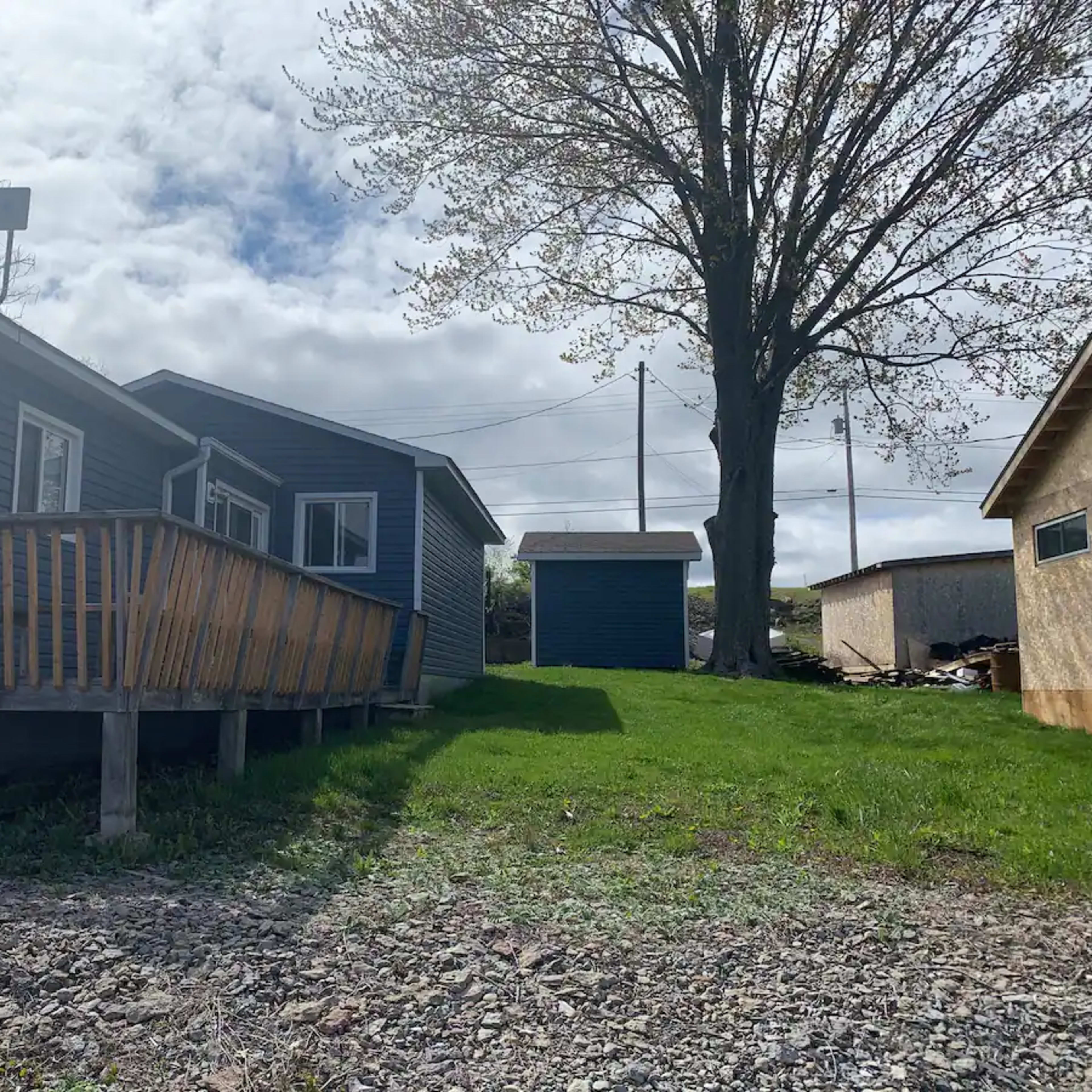 A blue house with a wooden deck is situated next to a small shed on a grassy yard, under a cloudy sky.