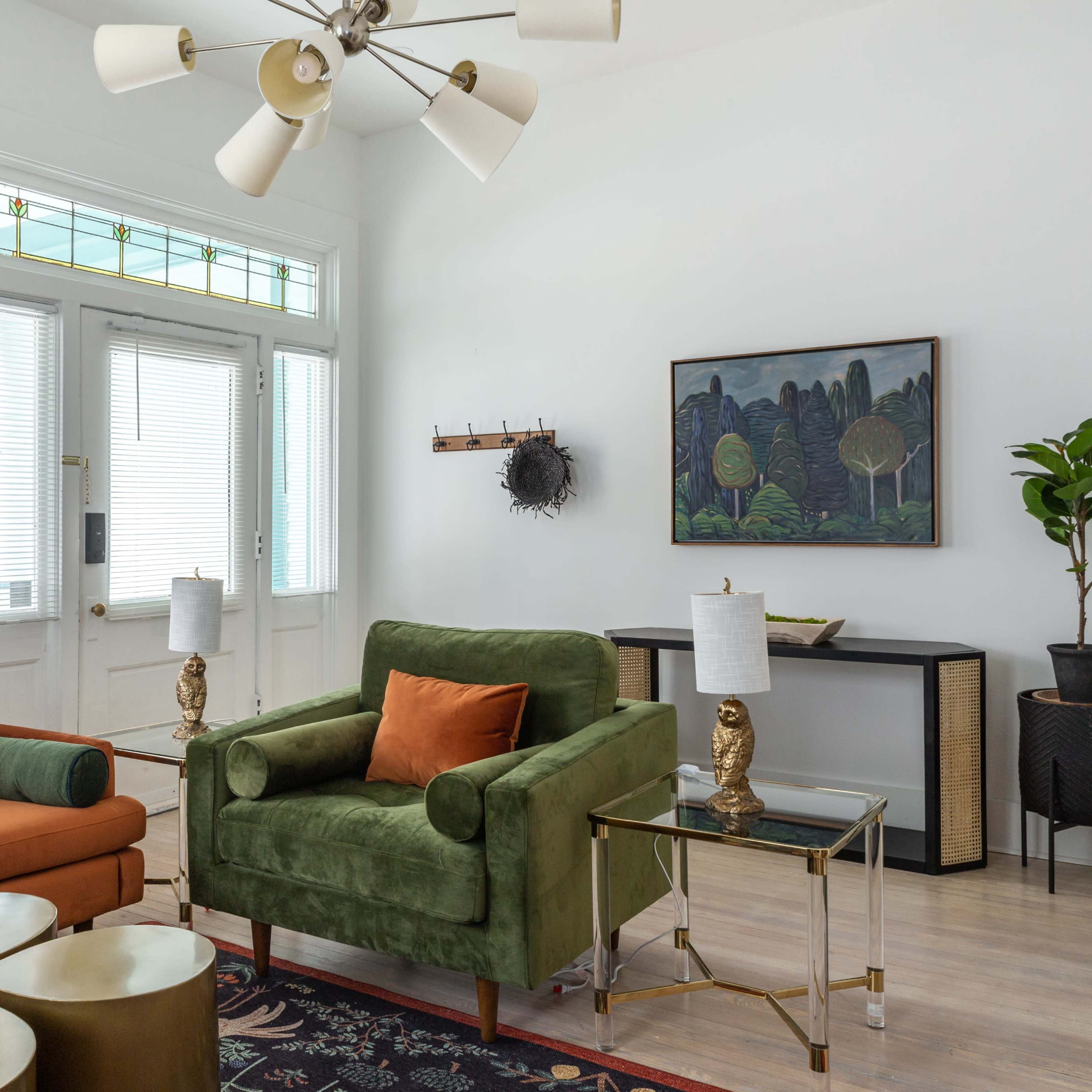 The living room features a green armchair and an orange sofa, with a coffee table, lamps, and artwork, set against a backdrop of large windows and light-colored walls.