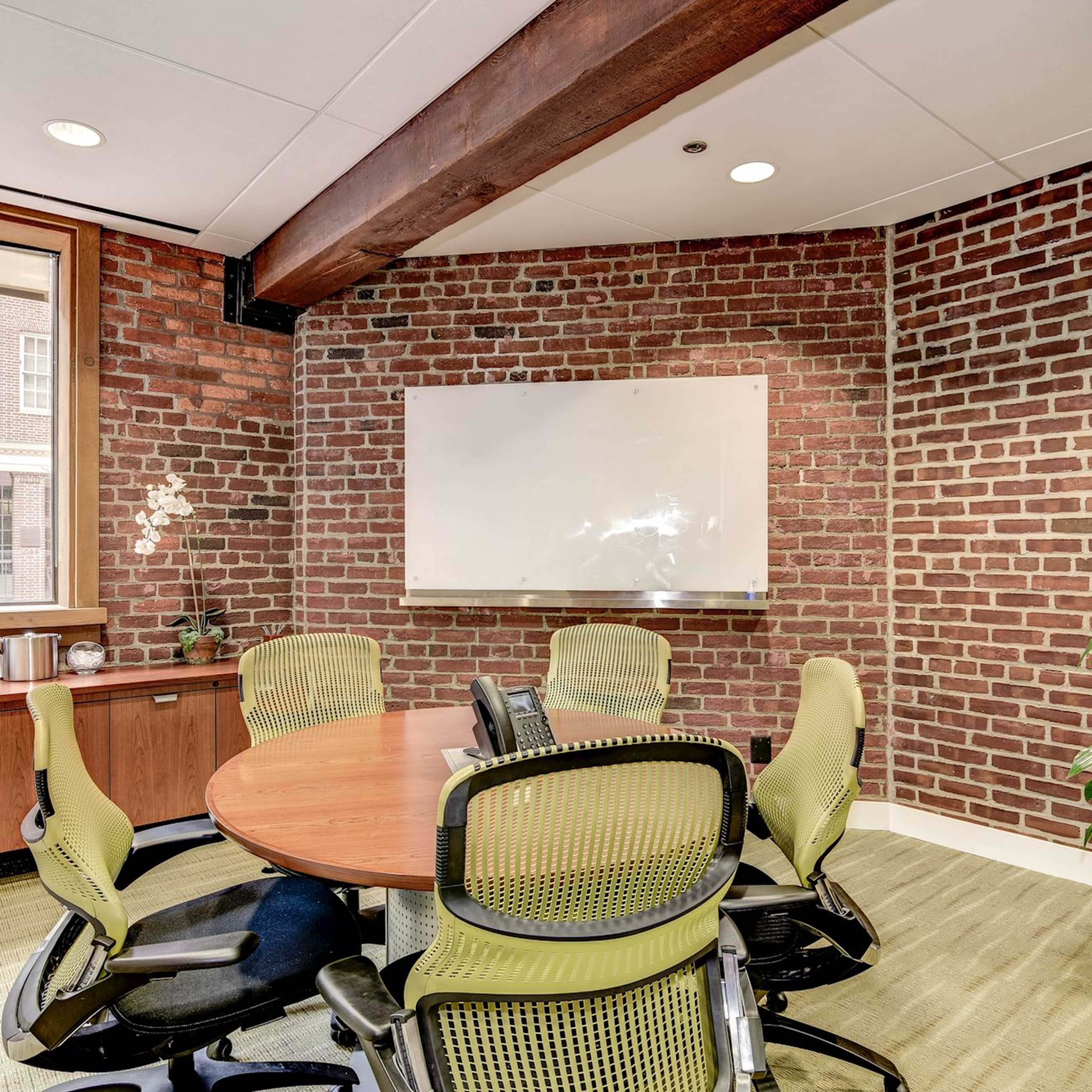 A conference room features a round table surrounded by green office chairs, a whiteboard on the wall, and a window overlooking a brick exterior.