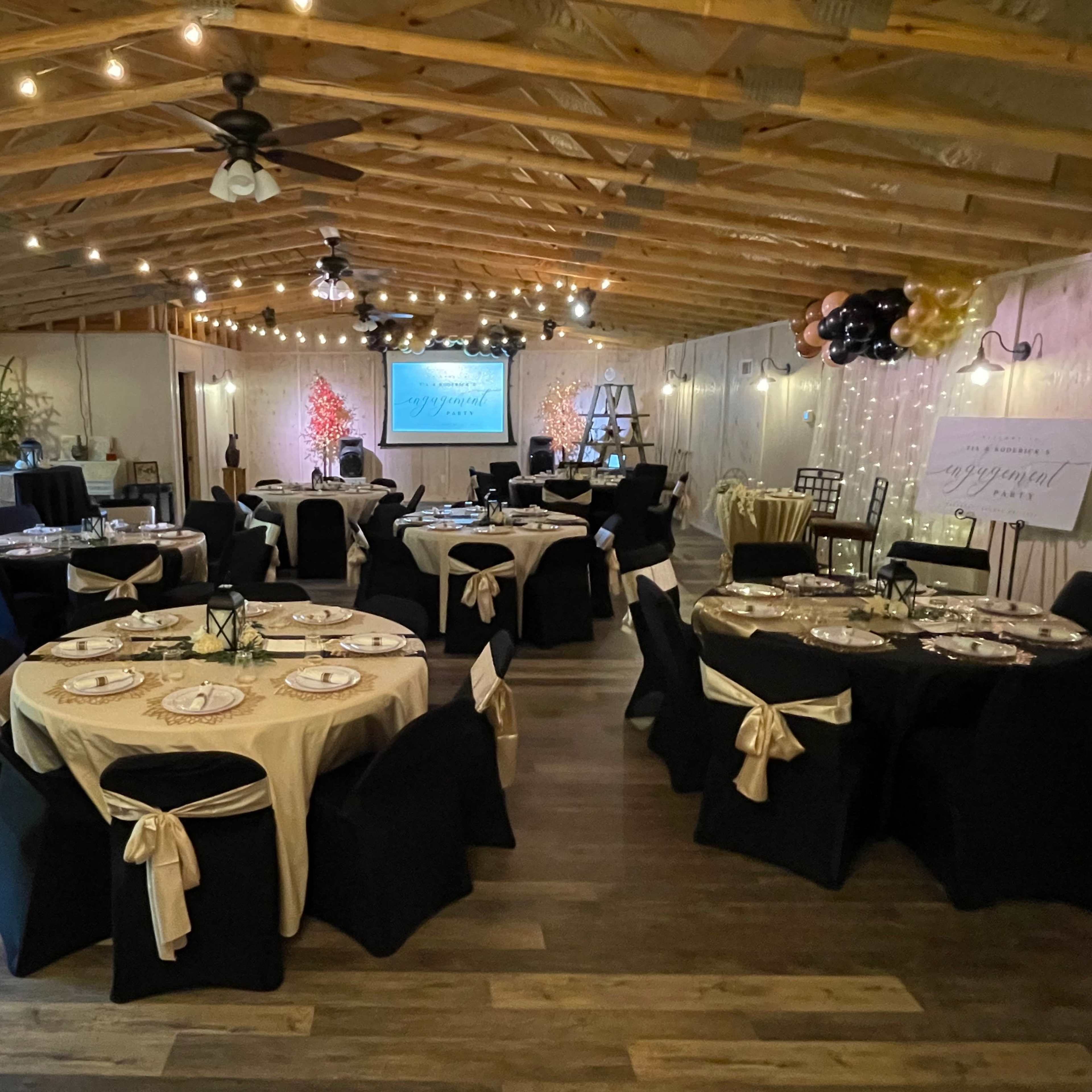 A decorated event space with multiple tables set for a formal gathering, featuring black and gold tablecloths and string lights hanging from the ceiling.