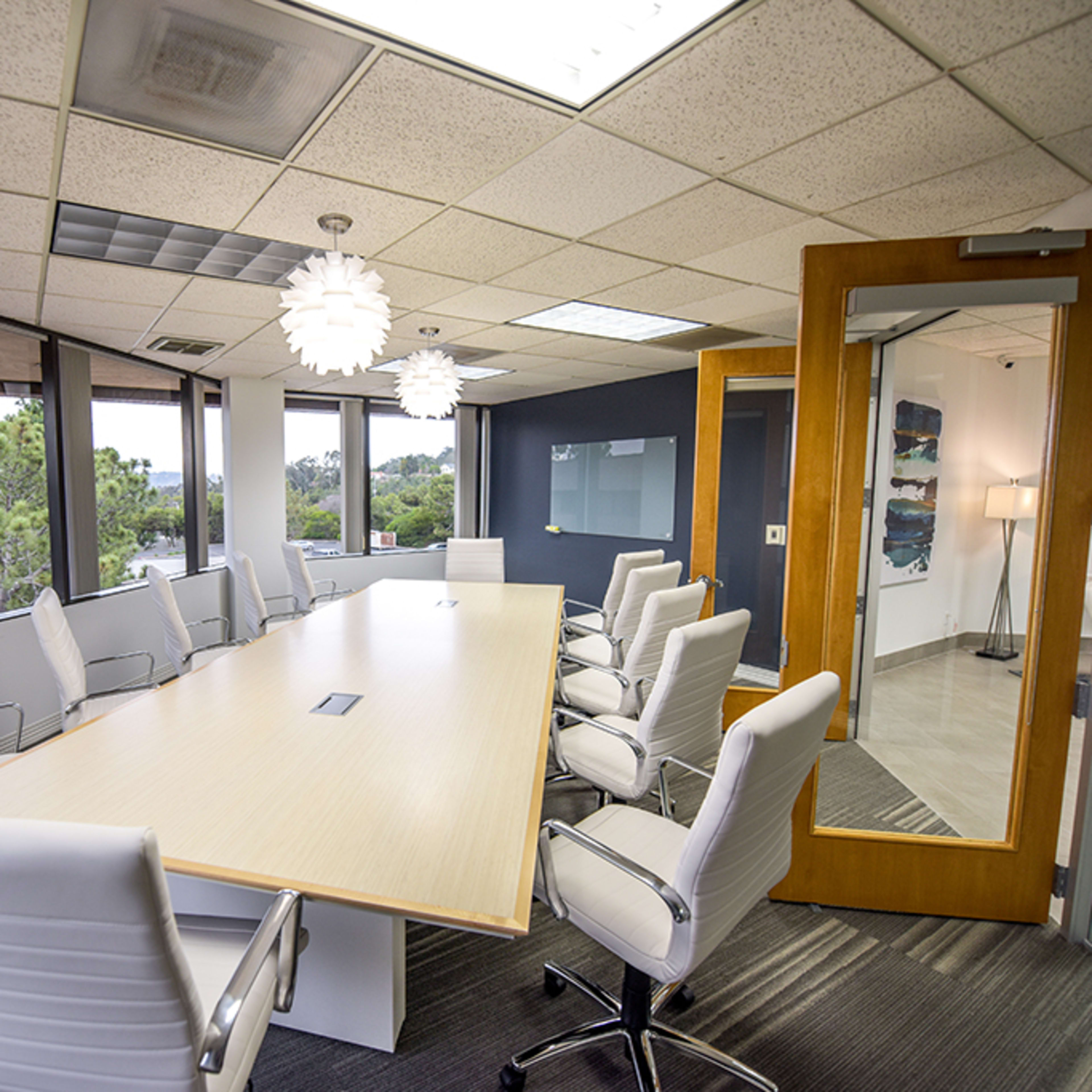 A modern conference room features a long wooden table surrounded by white chairs and large windows allowing natural light.