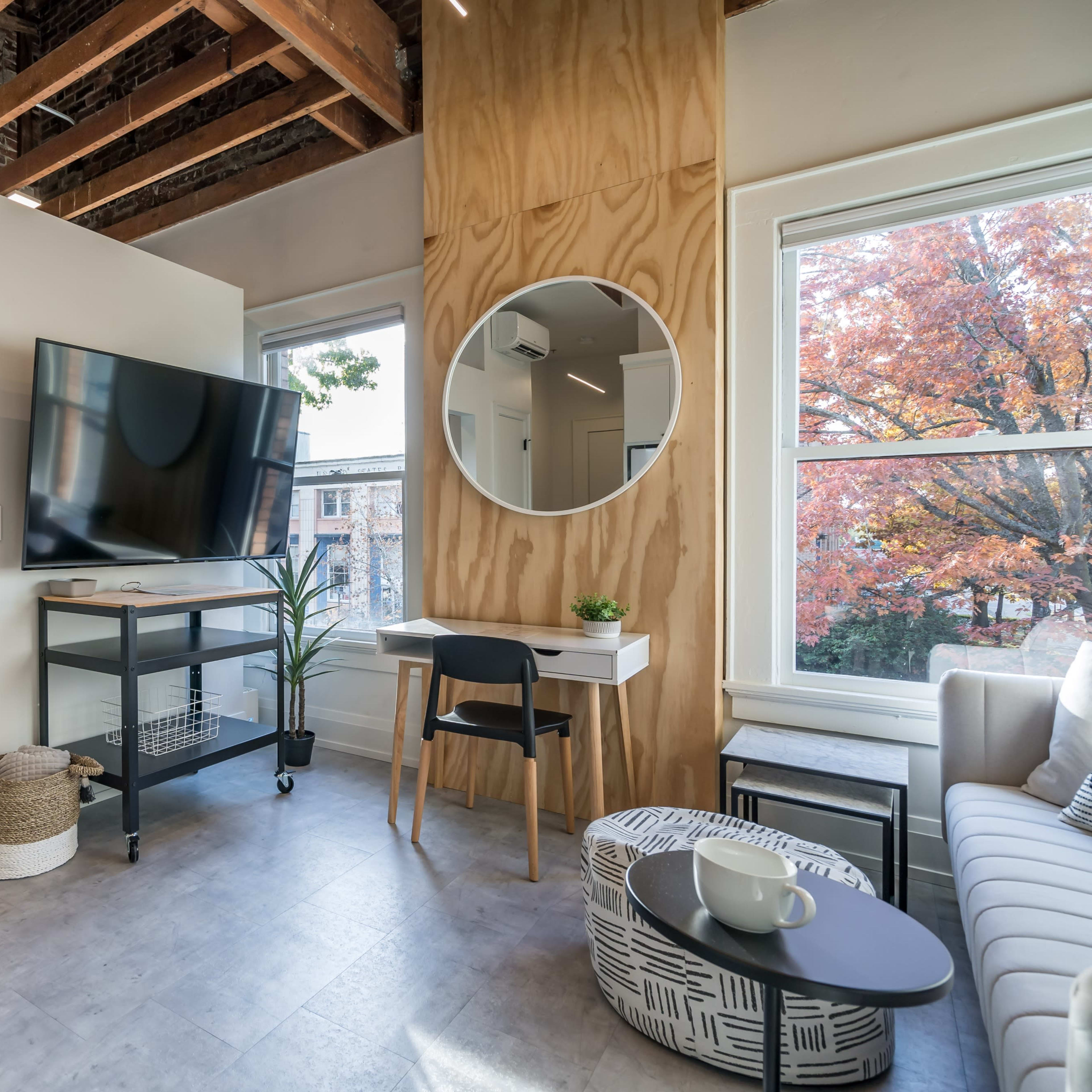 A modern studio apartment featuring a television mounted on a wooden wall, a round mirror, a small desk and chair, and a view of an outdoor autumn scene through a window.