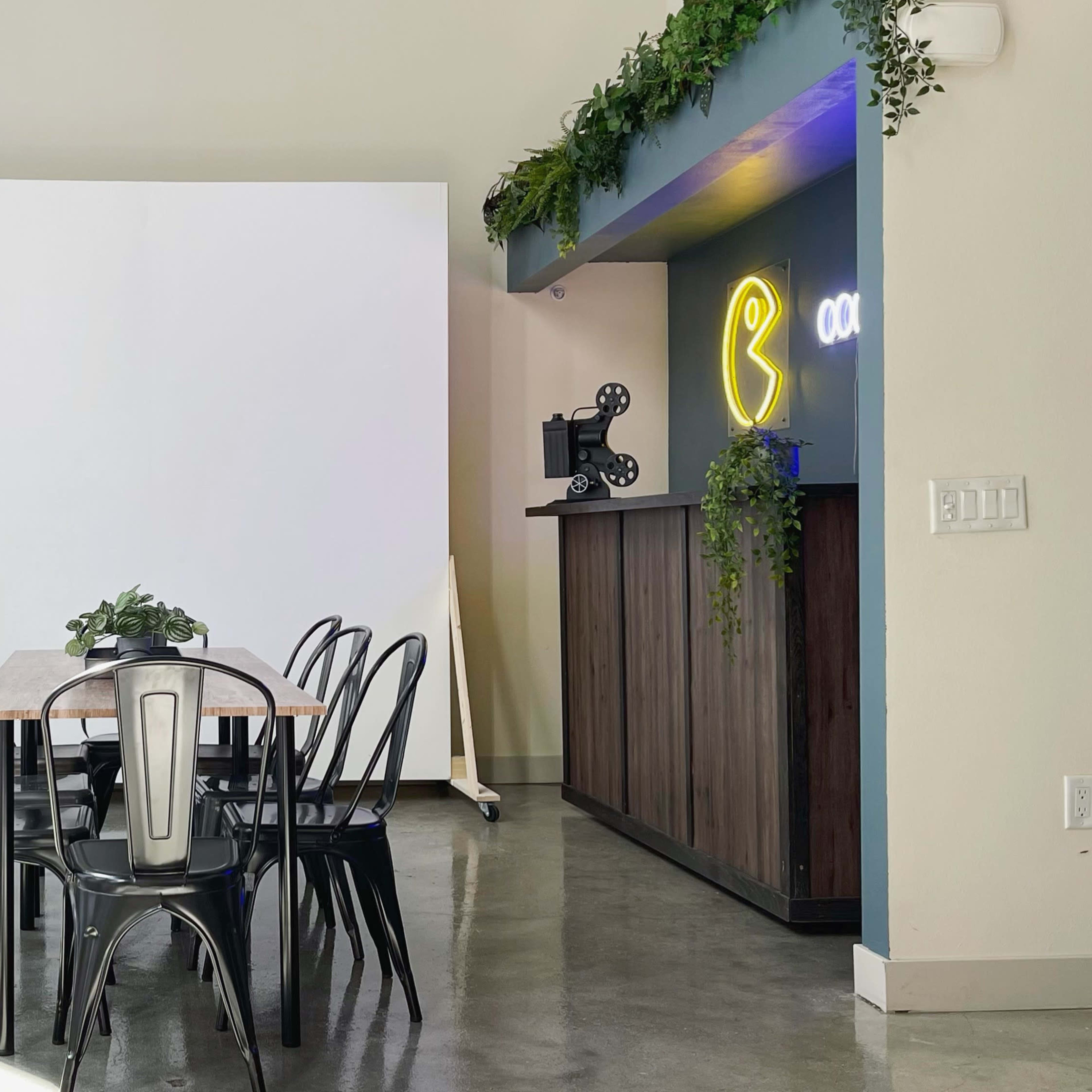 A modern interior space featuring a dining table surrounded by black chairs, a large blank white wall, and a bar area with neon lights and plants.