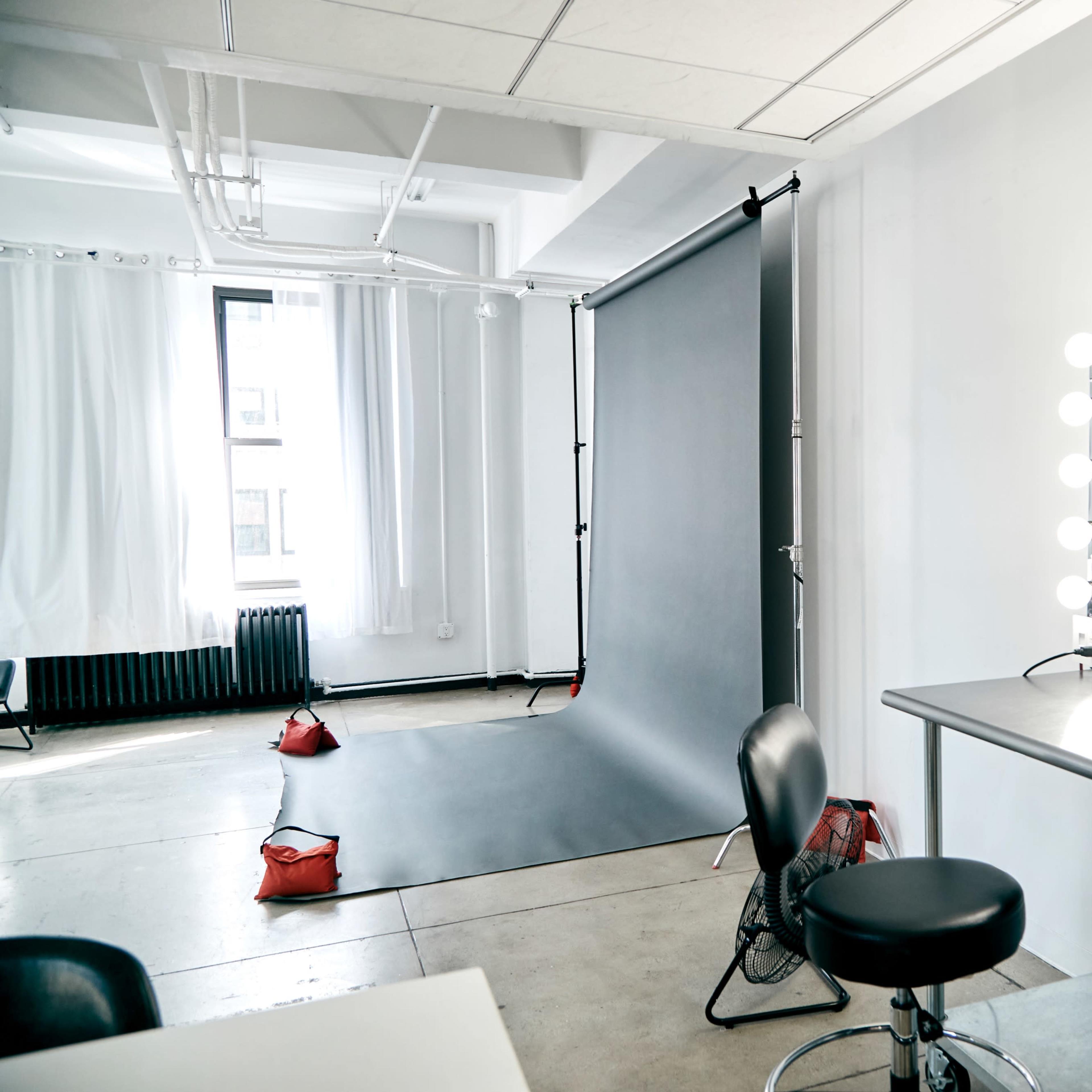 A photography studio featuring a black backdrop, a vanity mirror with lights, and a stool beside a metal table.