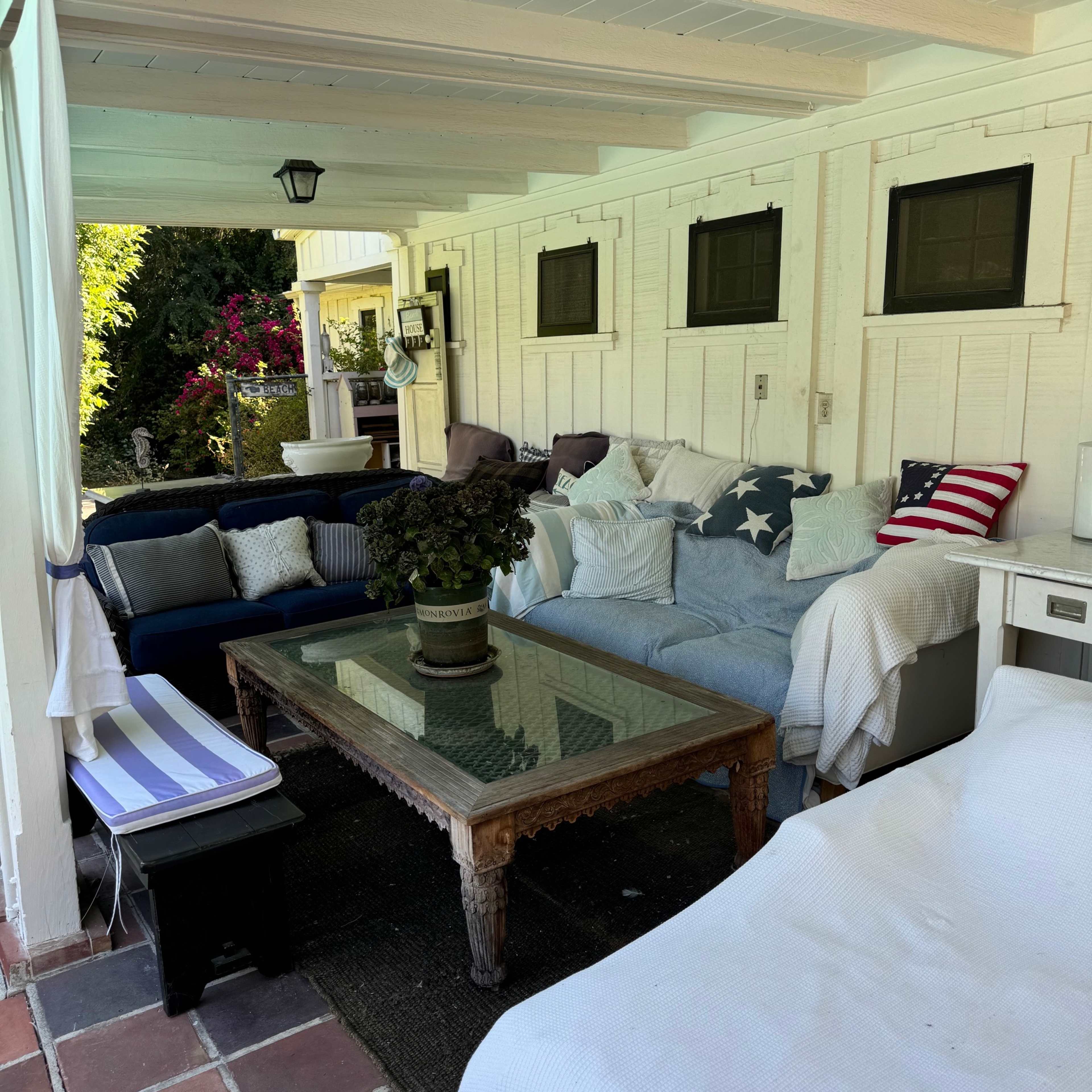 A covered patio area with several couches, a glass table in the center, and decorative plants.