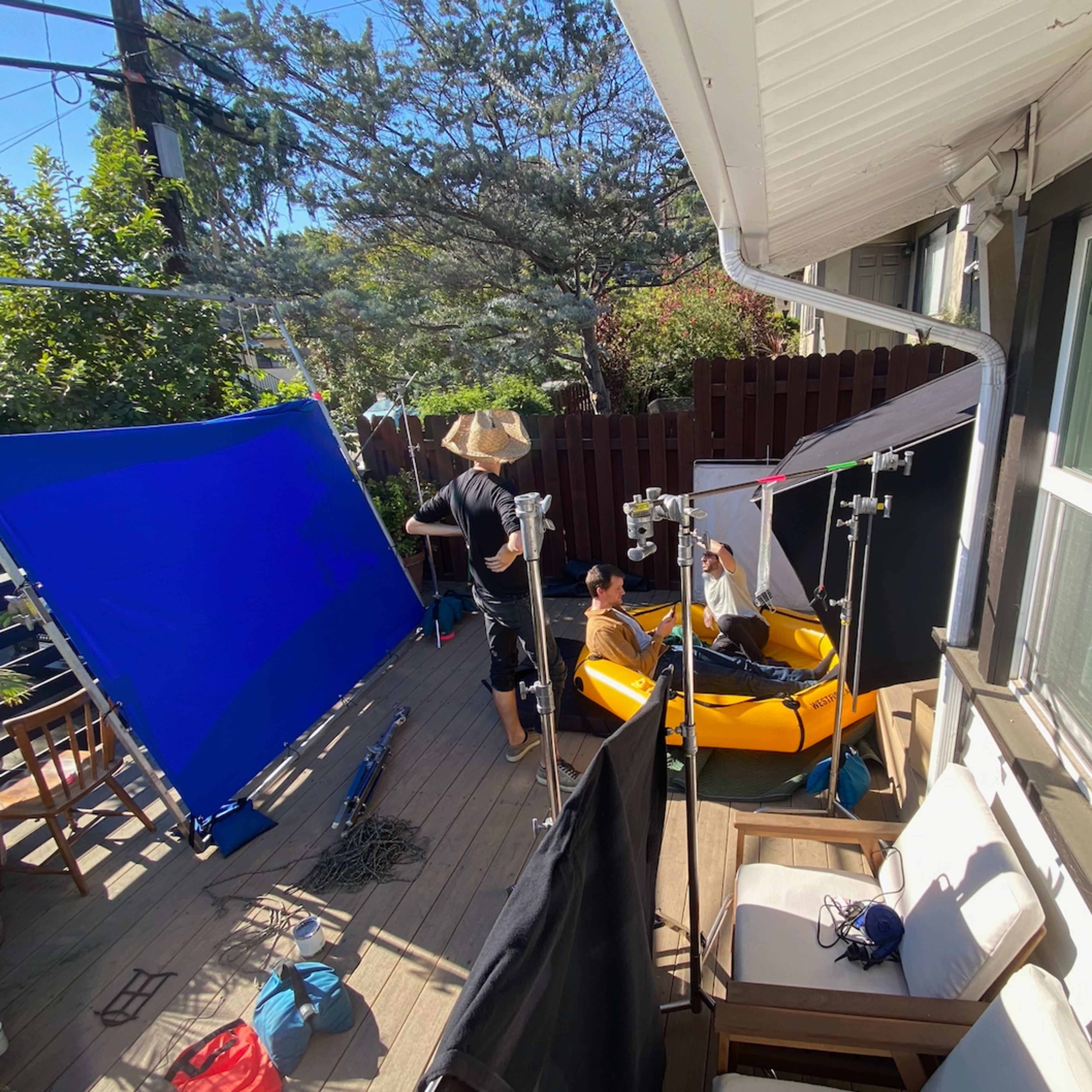 A film crew sets up lighting and backdrop equipment on a deck while a person sits in a yellow kayak.