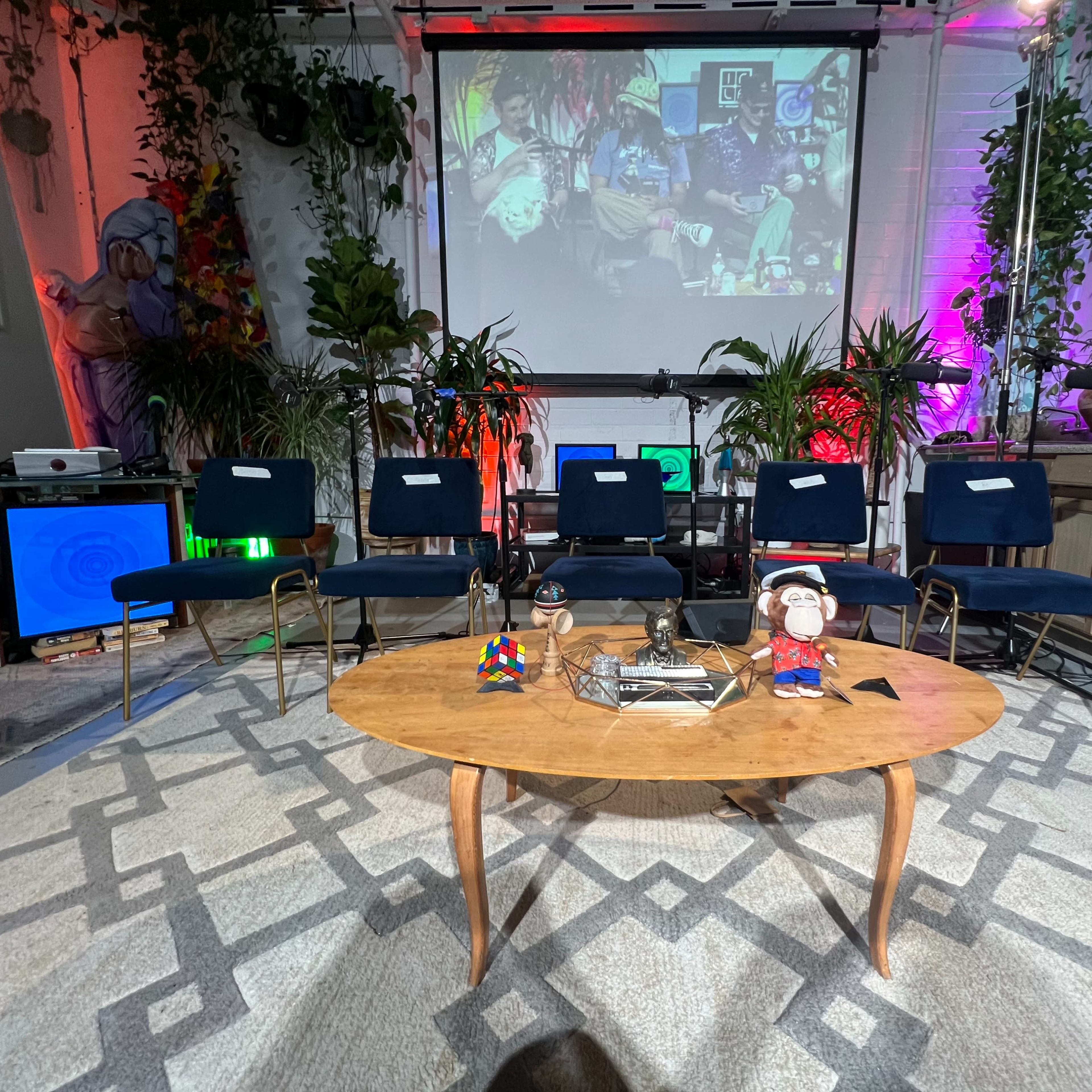 A set up for an event with a coffee table and several chairs arranged in front of a large screen displaying colorful artwork, surrounded by plants and decorative elements.
