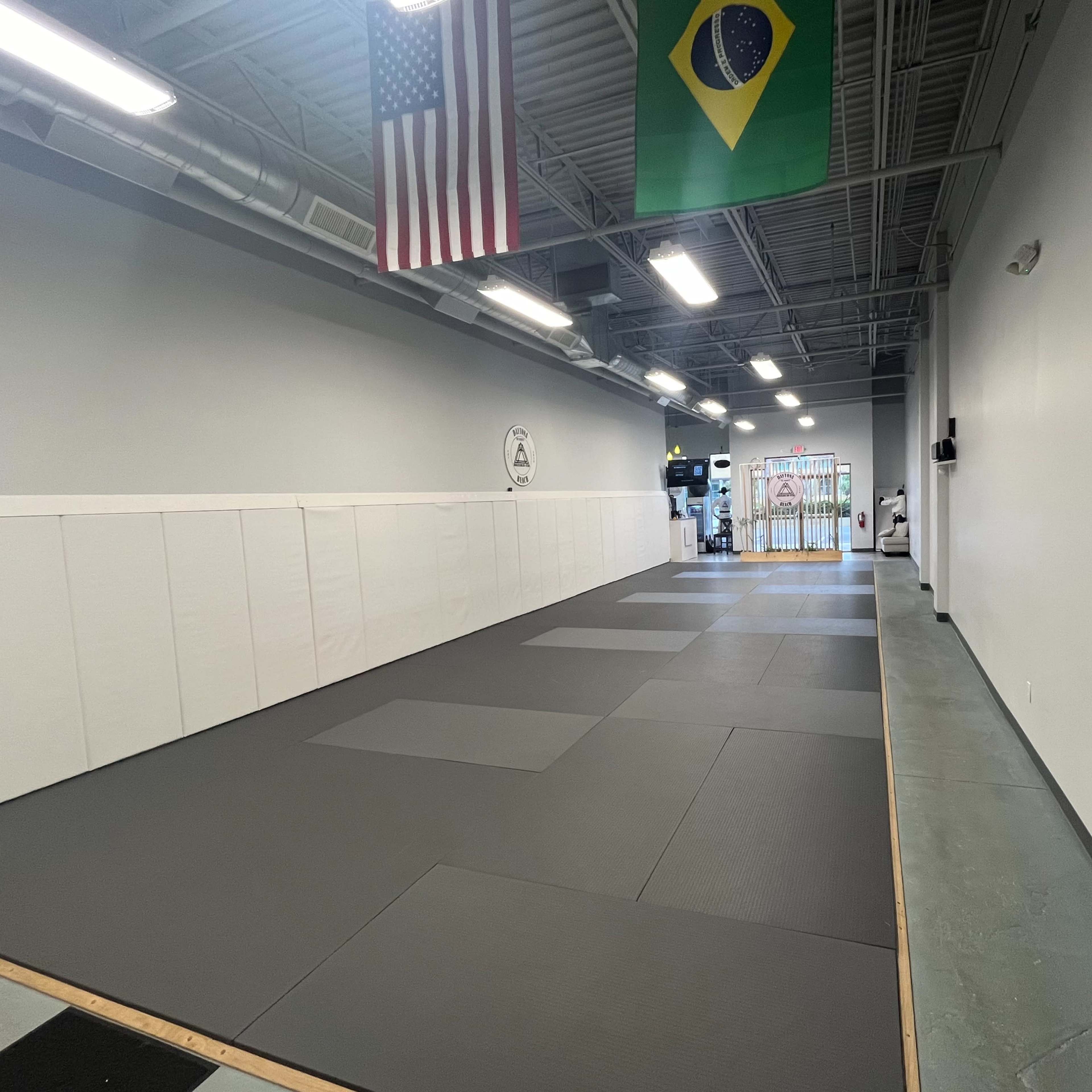 An empty martial arts training room featuring gray mats, white wall padding, and flags of the United States and Brazil hanging from the ceiling.