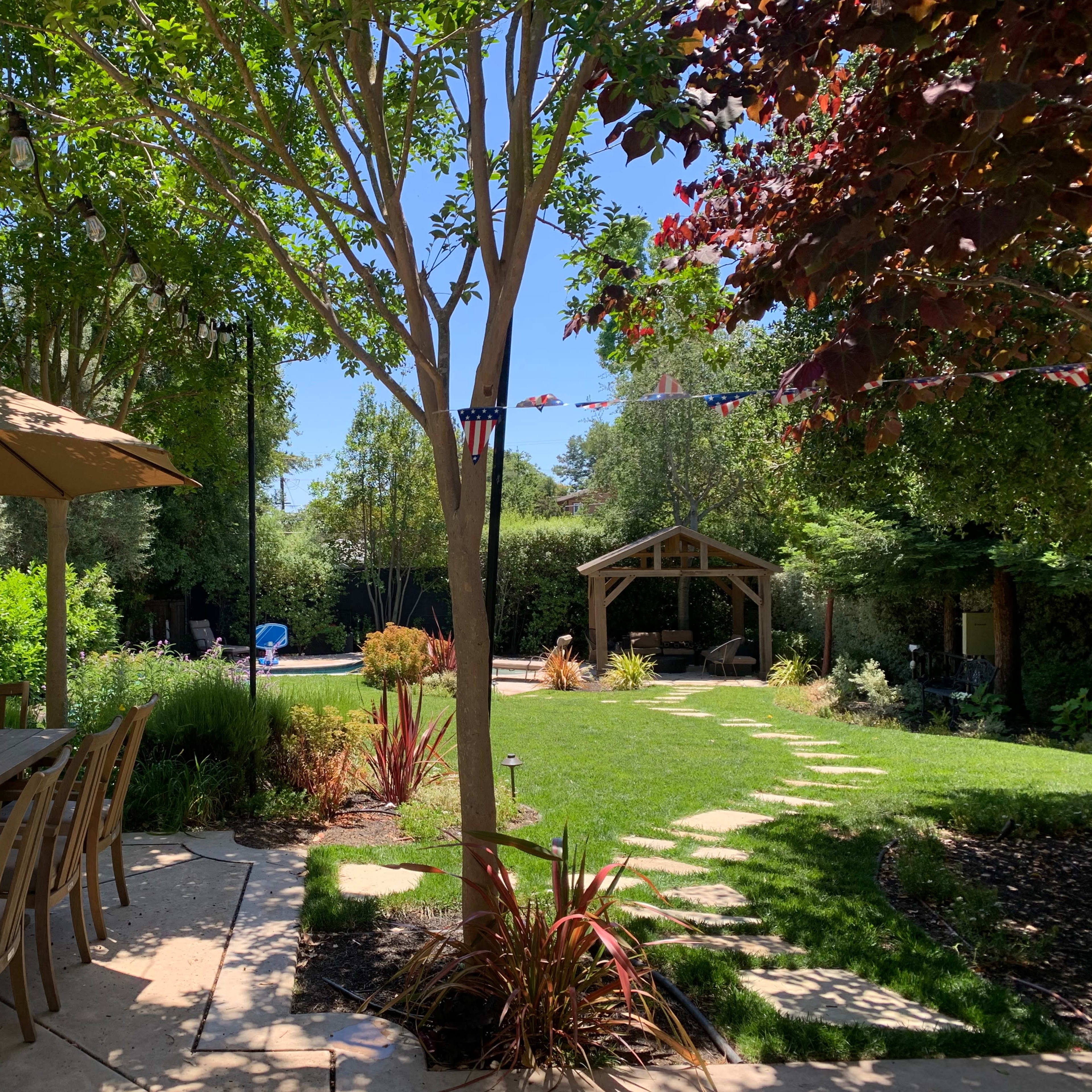 A patio area with a wooden dining table and umbrella overlooks a landscaped garden featuring a lawn, stone pathway, and a gazebo.
