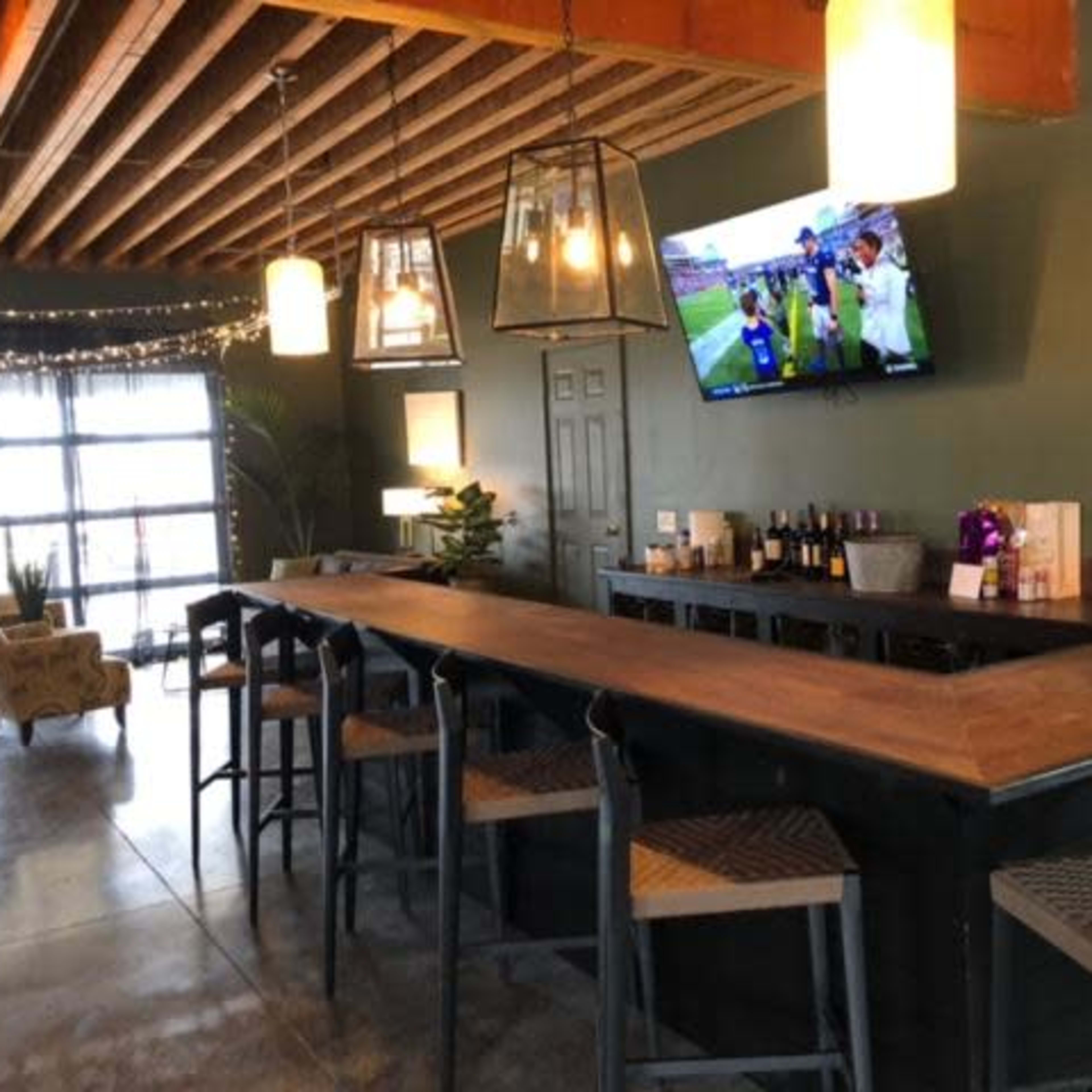 A modern bar area features a long wooden countertop, high stools, and a television displaying a sports event, with soft lighting and a lounge area in the background.