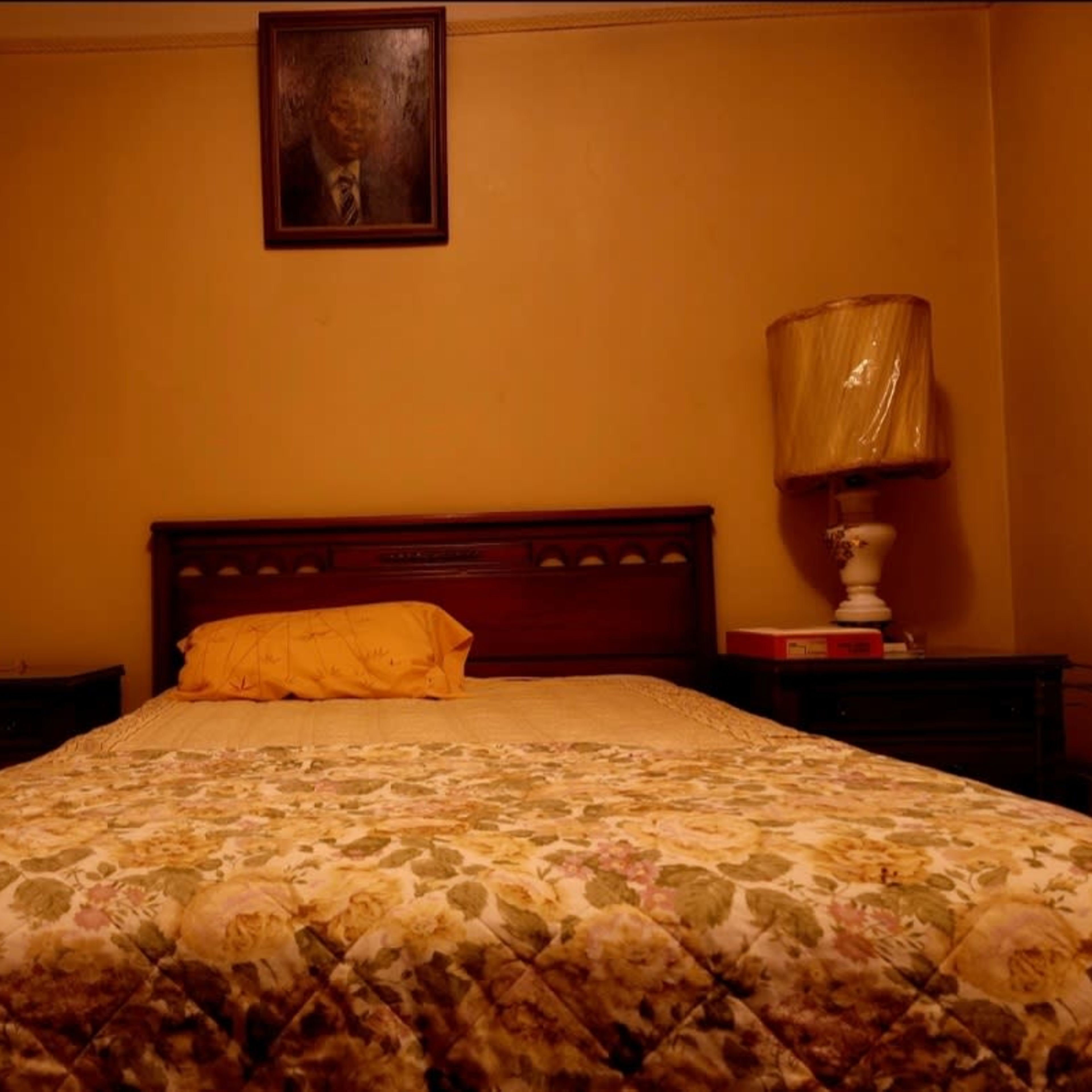 A simple bedroom with a double bed, a floral patterned blanket, bedside tables, a lamp, and a portrait hanging on the wall above the bed.