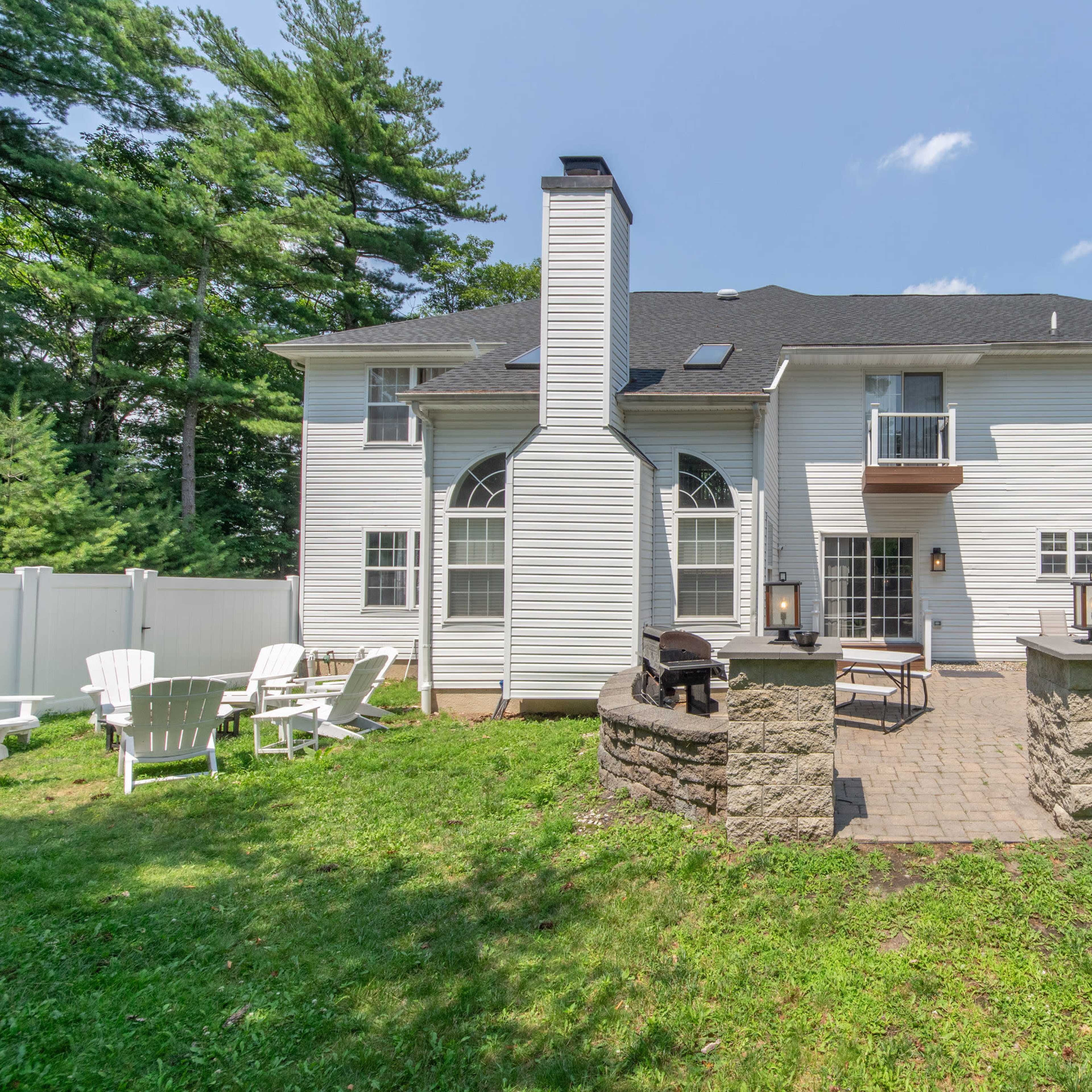 The backyard of a two-story house with a stone patio, outdoor seating, and a grassy area surrounded by trees.