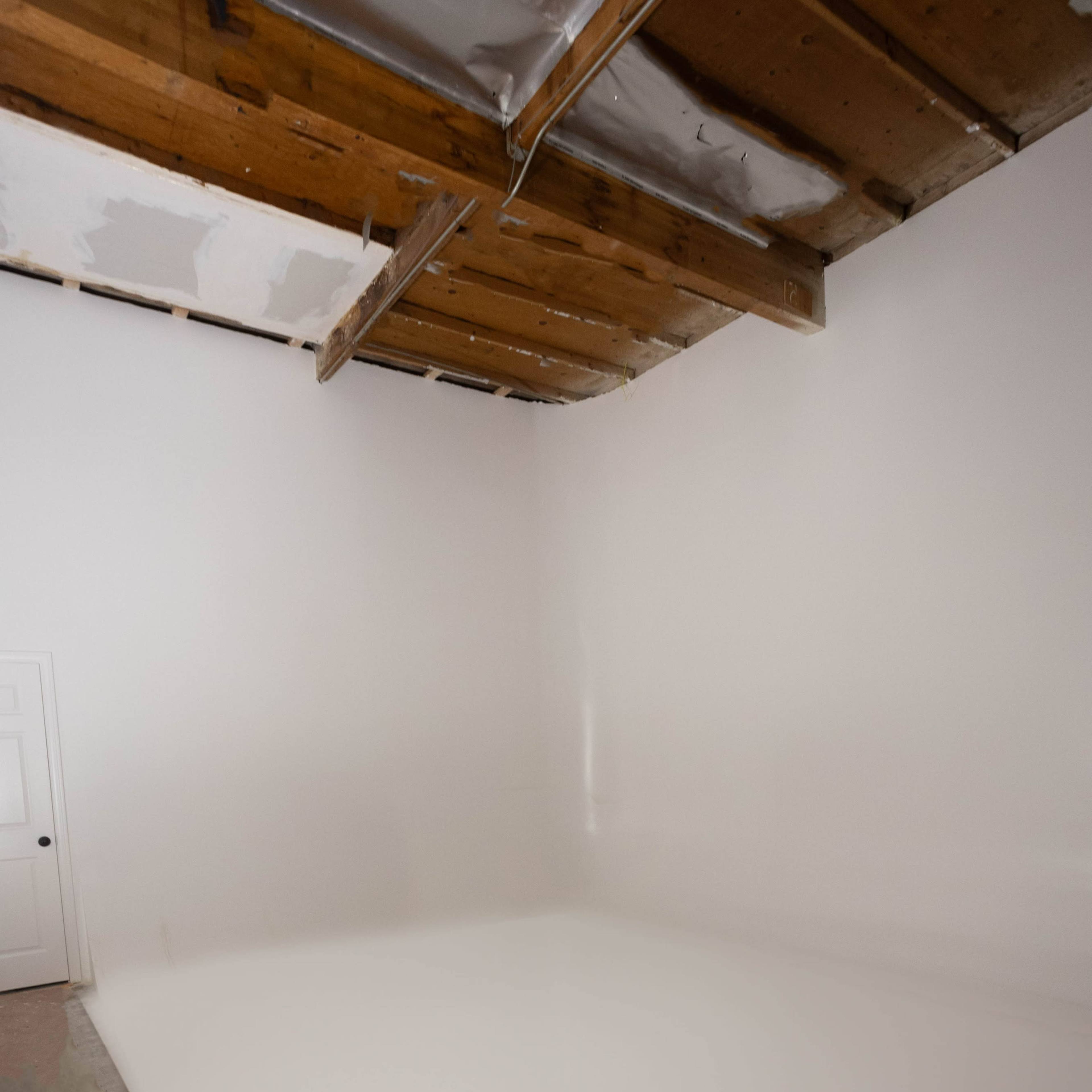 An empty room with a smooth white floor and white walls, featuring exposed wooden beams on the ceiling and a closed door.