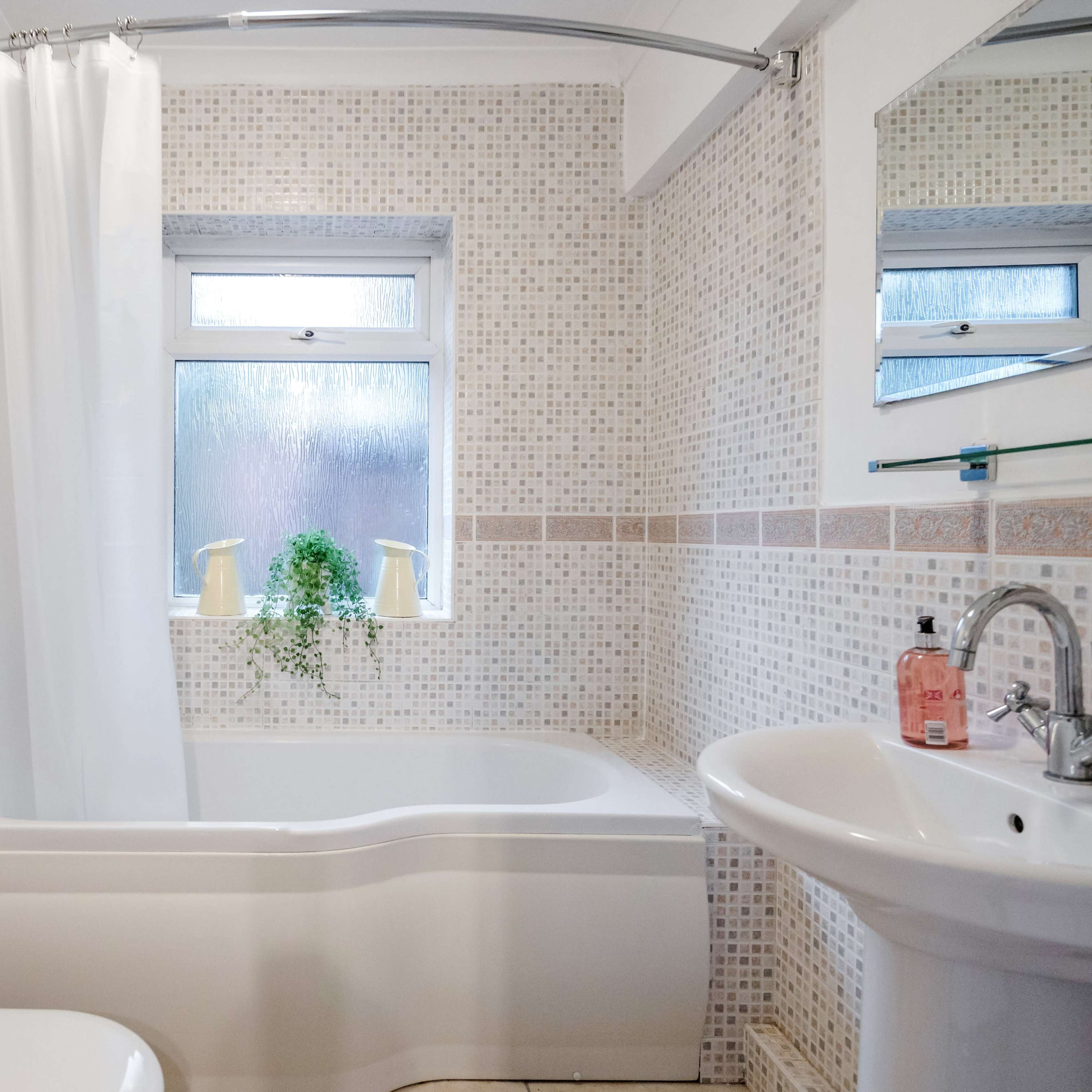 A bathroom with a curved bathtub, a shower curtain, a window with plants, and a sink against tiled walls.
