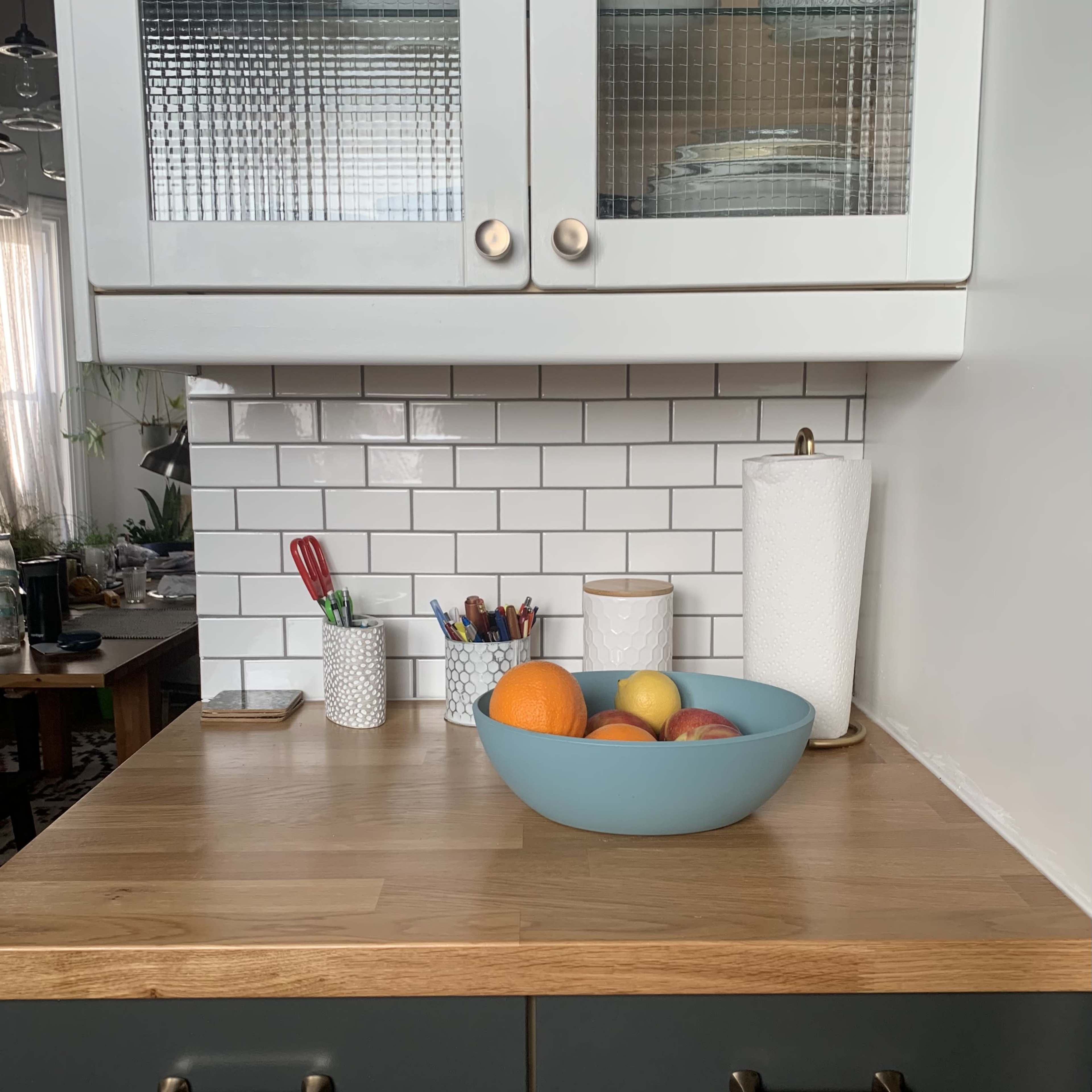 A countertop displays a blue bowl of assorted fruits, surrounded by utensils and containers on a kitchen surface.