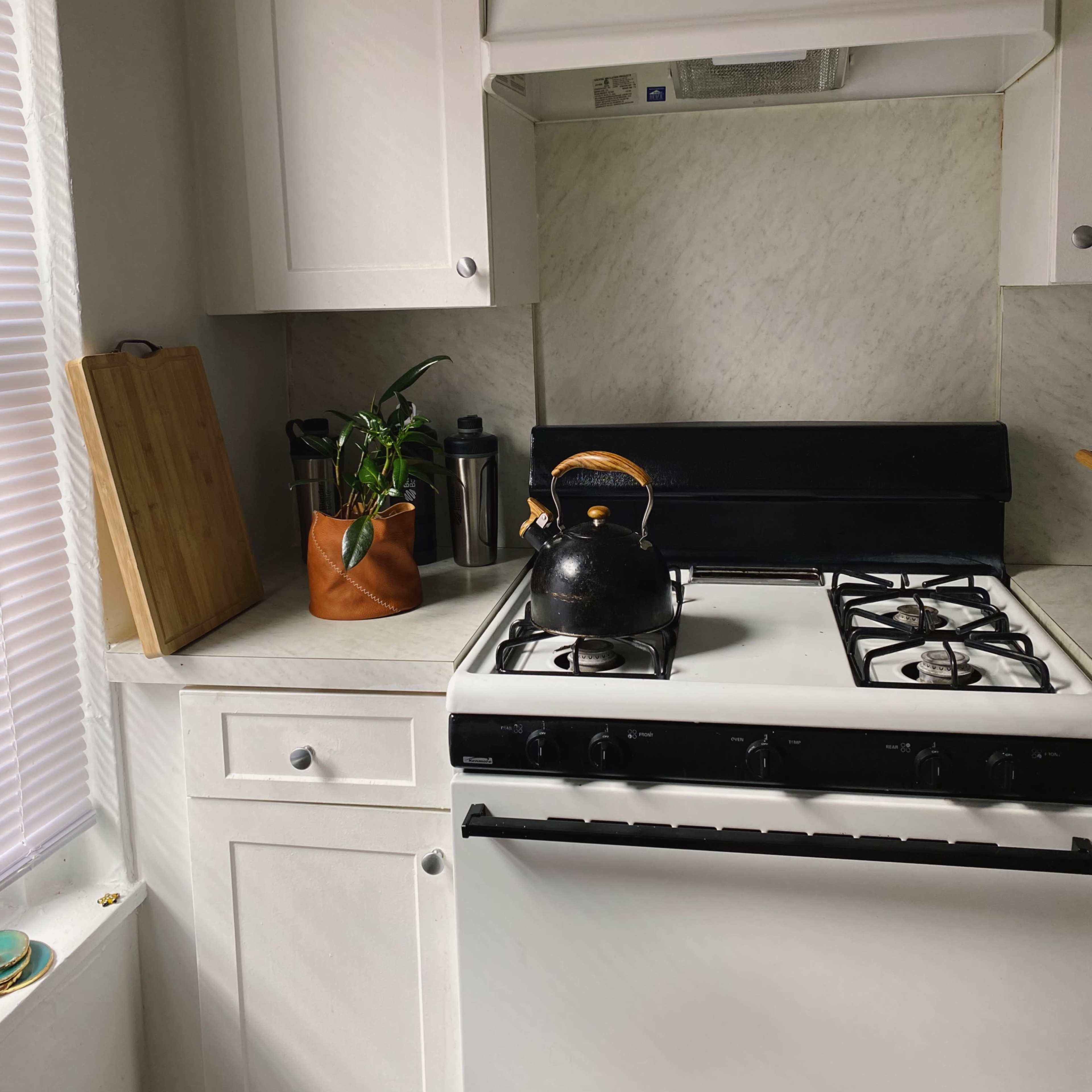 A black kettle sits on a gas stove in a modern kitchen with white cabinetry and a marble backsplash.