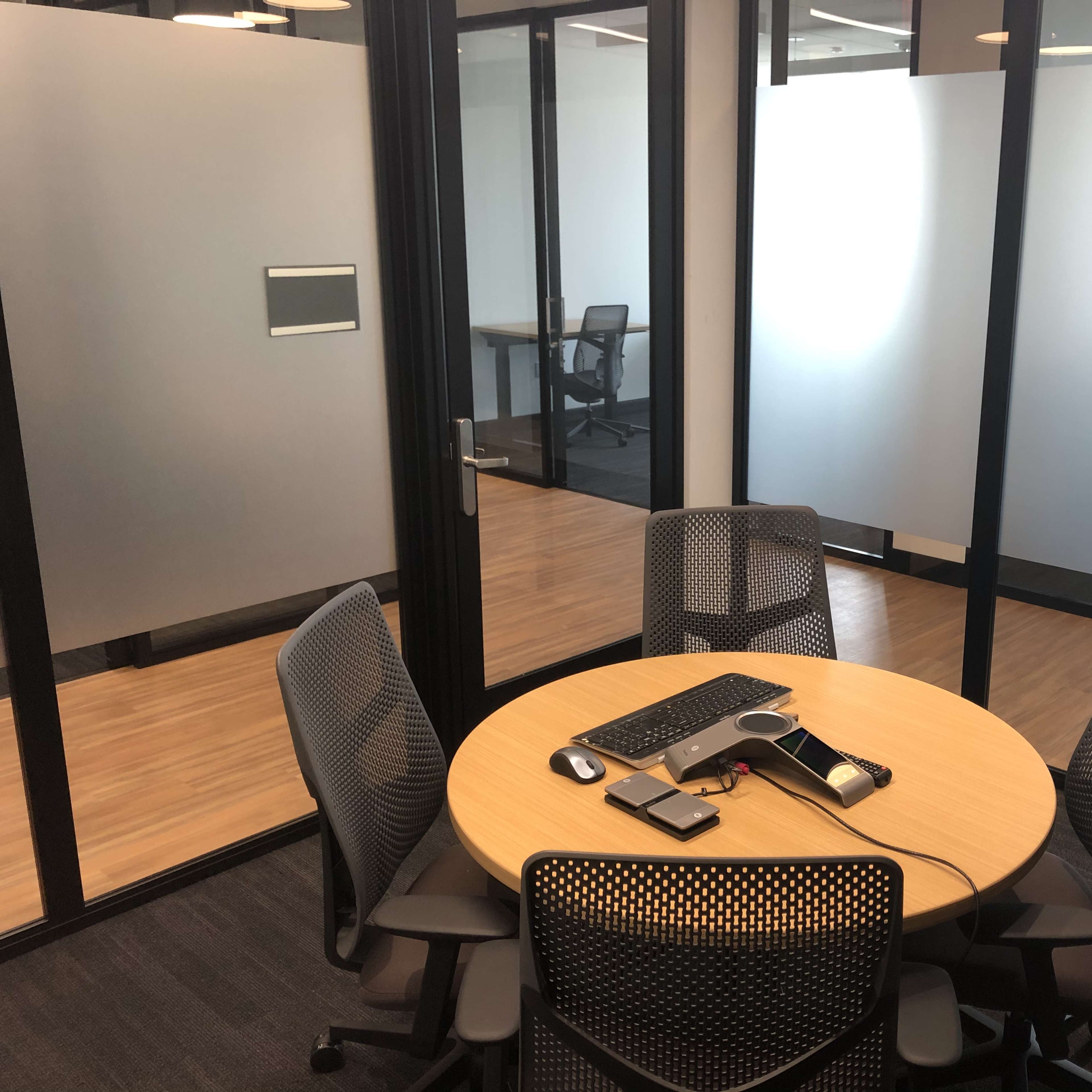 A round table with a computer and phone sits in a glass-walled meeting room featuring four mesh chairs and wooden flooring.