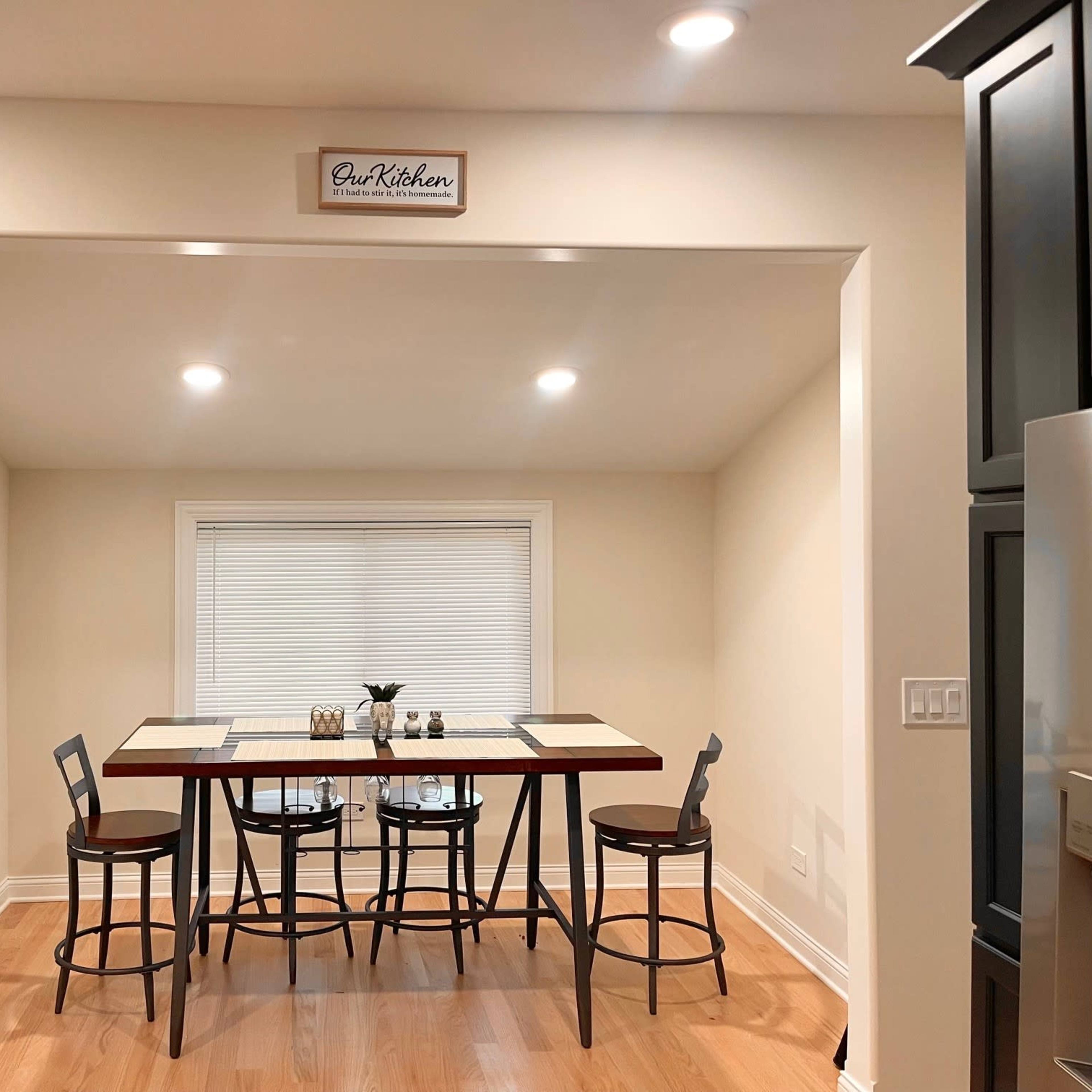 A small dining area features a table with four chairs and a window above, illuminated by overhead lights.