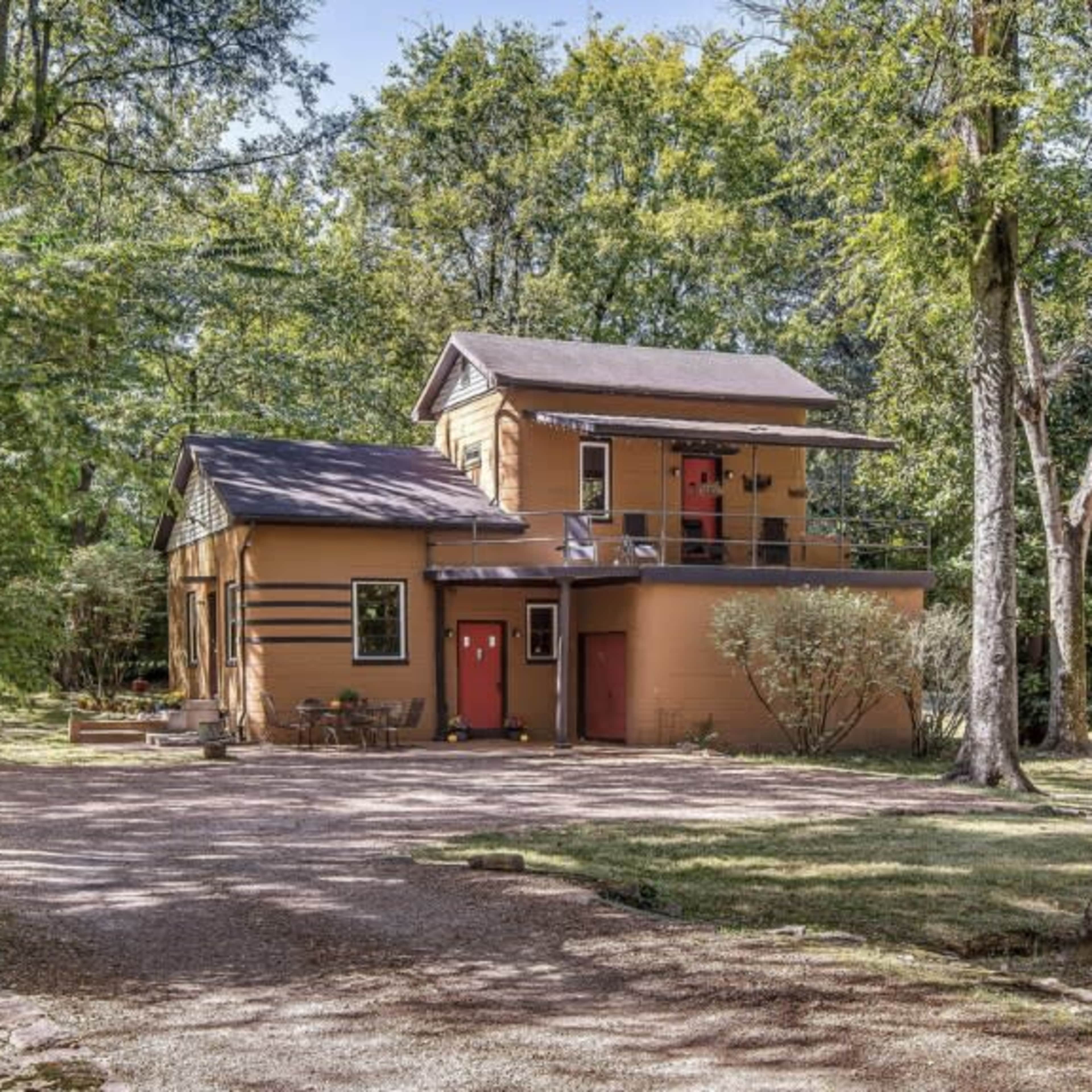 A two-story house with a balcony and red doors is surrounded by trees and gravel pathways.