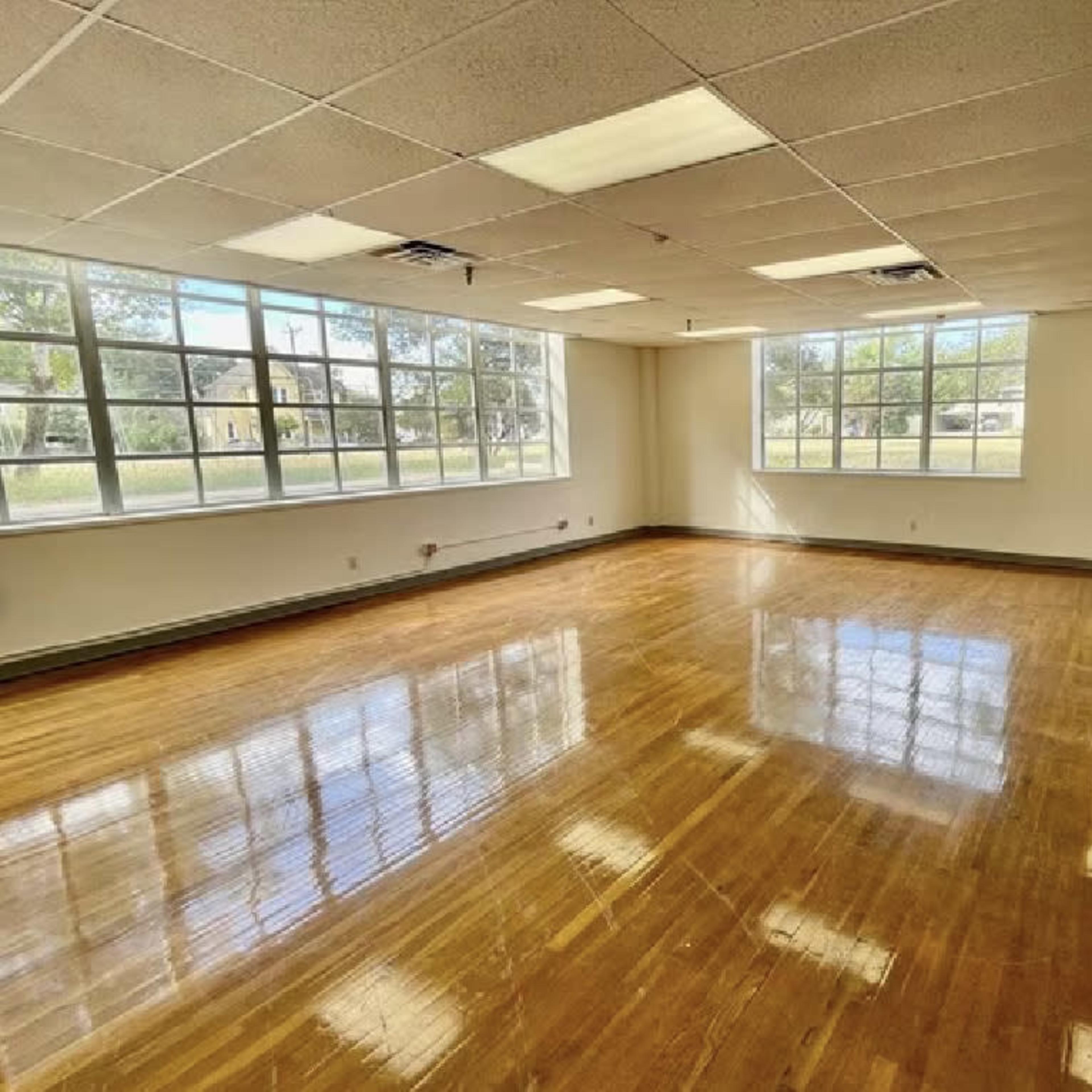 A vacant room with polished wooden floors and large windows allowing natural light to enter.