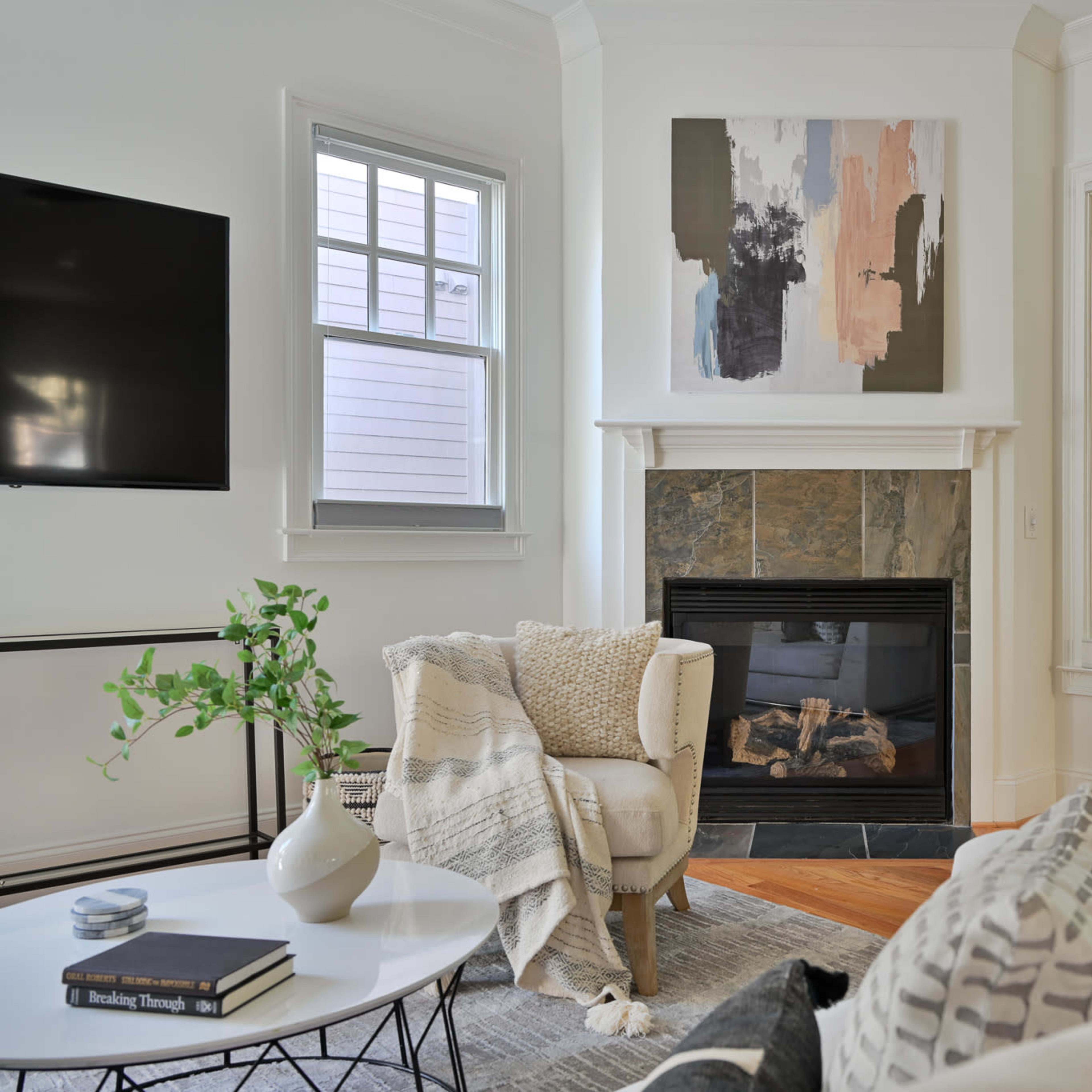 A bright living room featuring a fireplace, a large abstract painting on the wall, a coffee table with books, and large windows that overlook a green outdoor space.