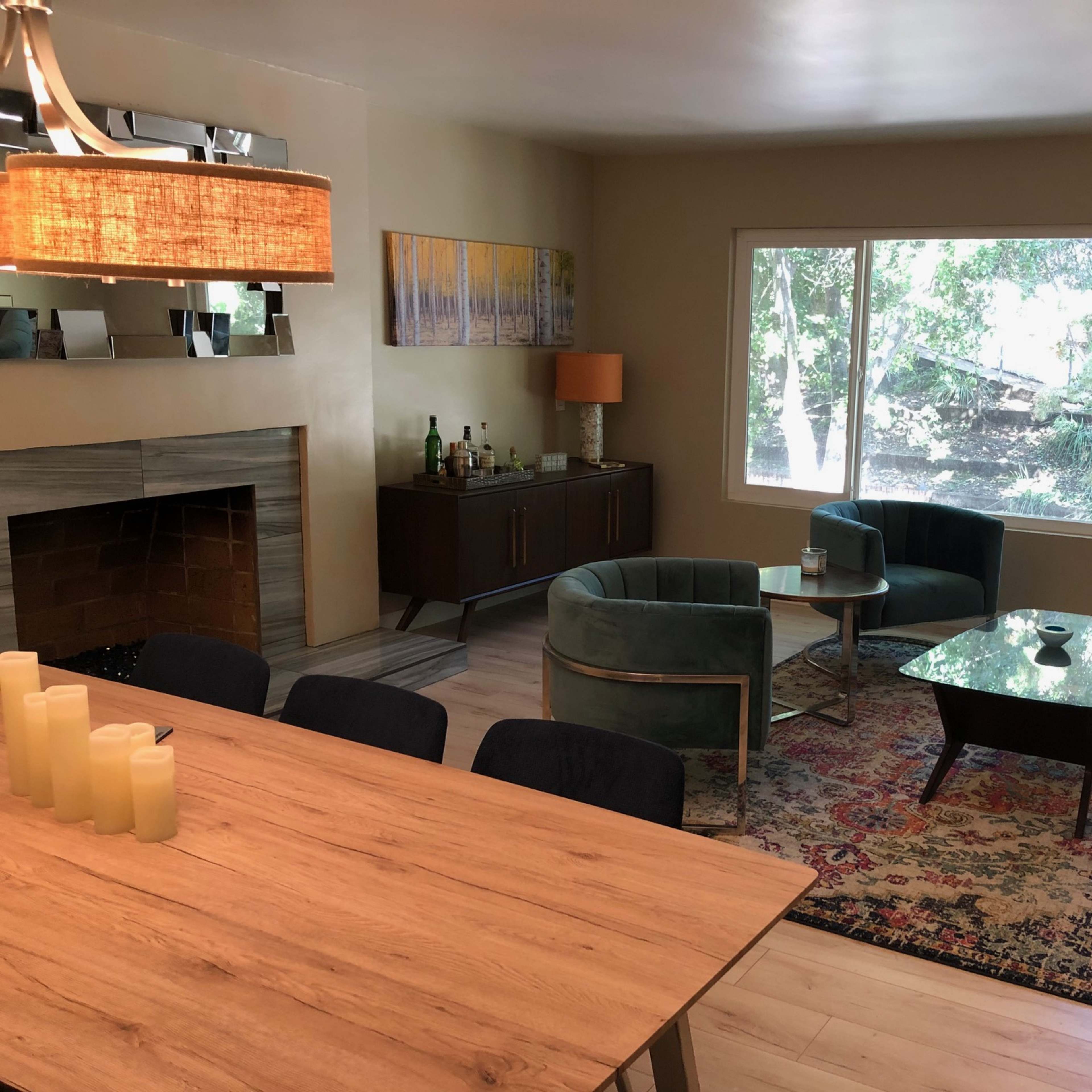 The room features a living area with a fireplace, two green chairs, a brown sofa, a coffee table, and a dining table, all arranged near large windows that provide natural light.