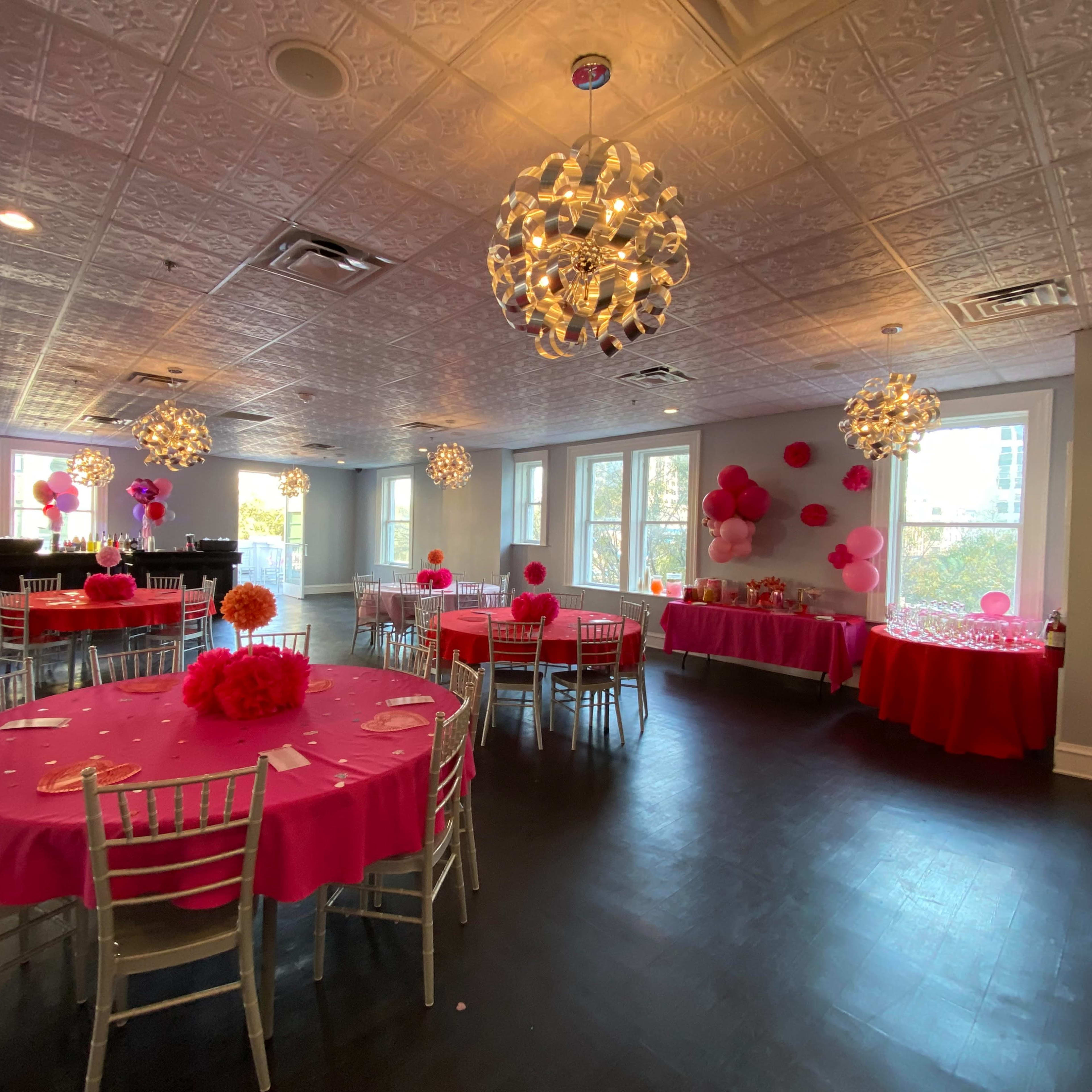 A spacious event room features tables decorated with pink tablecloths and centerpieces, illuminated by decorative chandeliers.