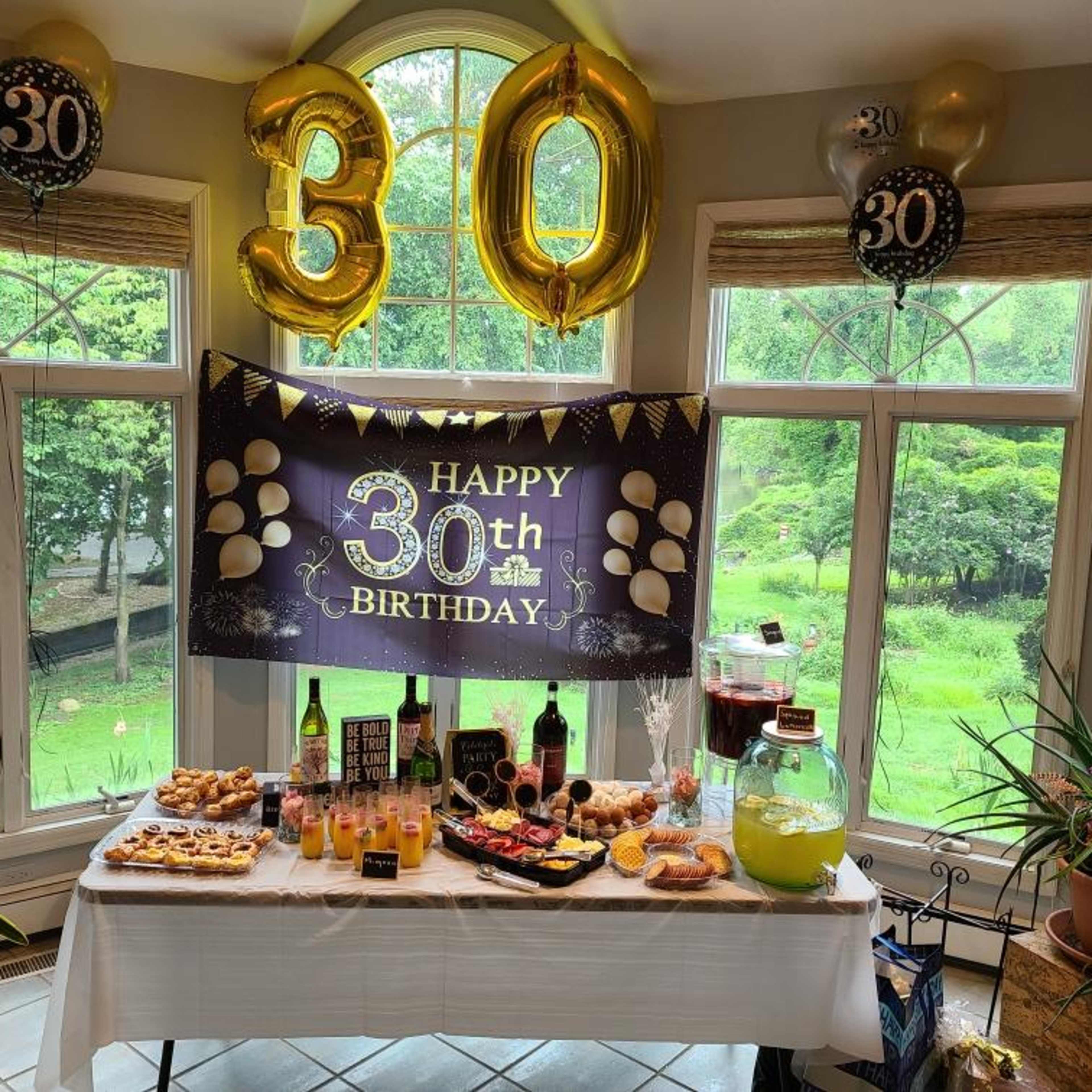 The scene features a decorated table filled with food and drinks for a 30th birthday celebration, accompanied by yellow balloon banners and a festive sign.