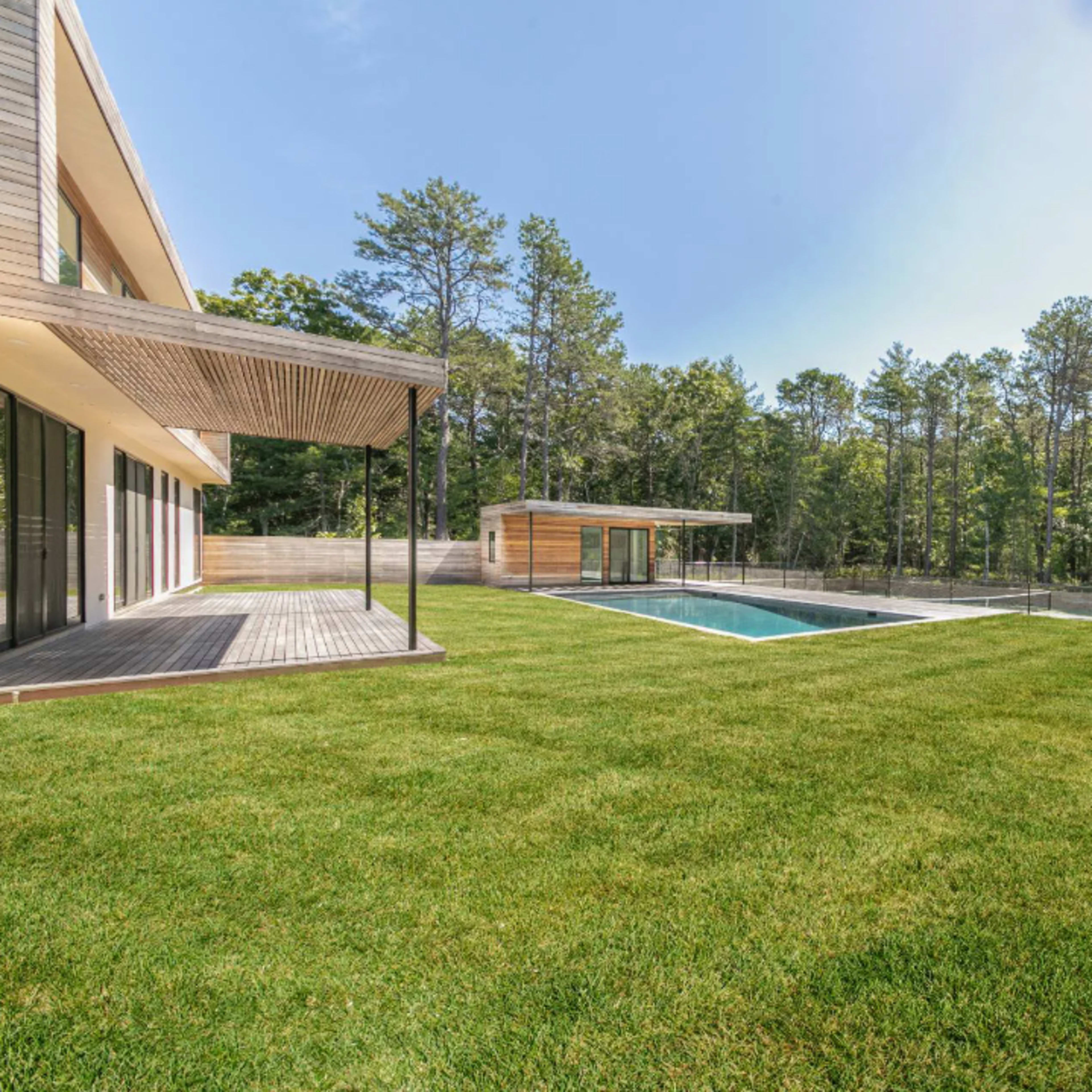 A modern house with large windows overlooks a rectangular swimming pool surrounded by a neatly manicured lawn and forested area.