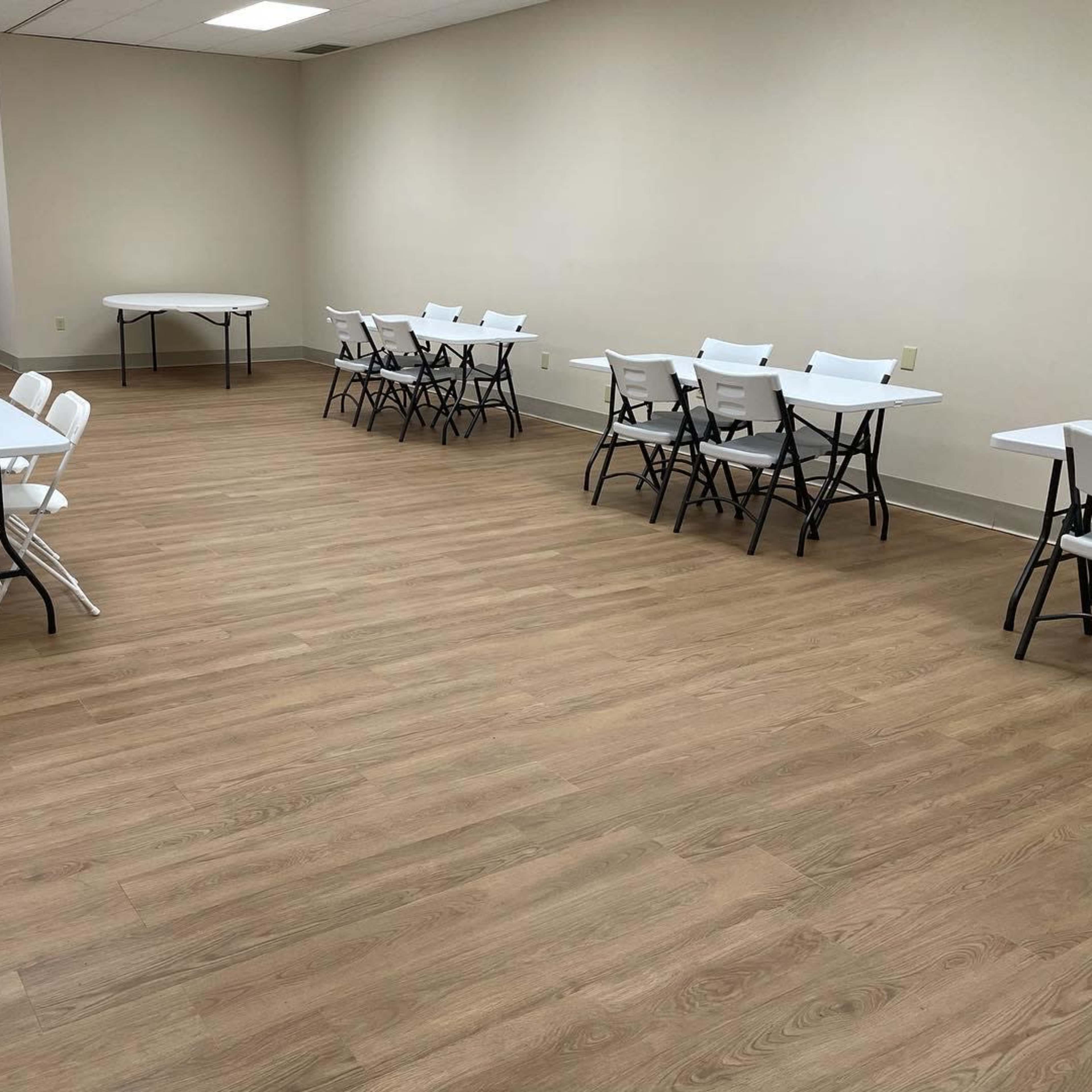 A bright, empty room with several white folding tables and chairs arranged neatly on a wooden floor.