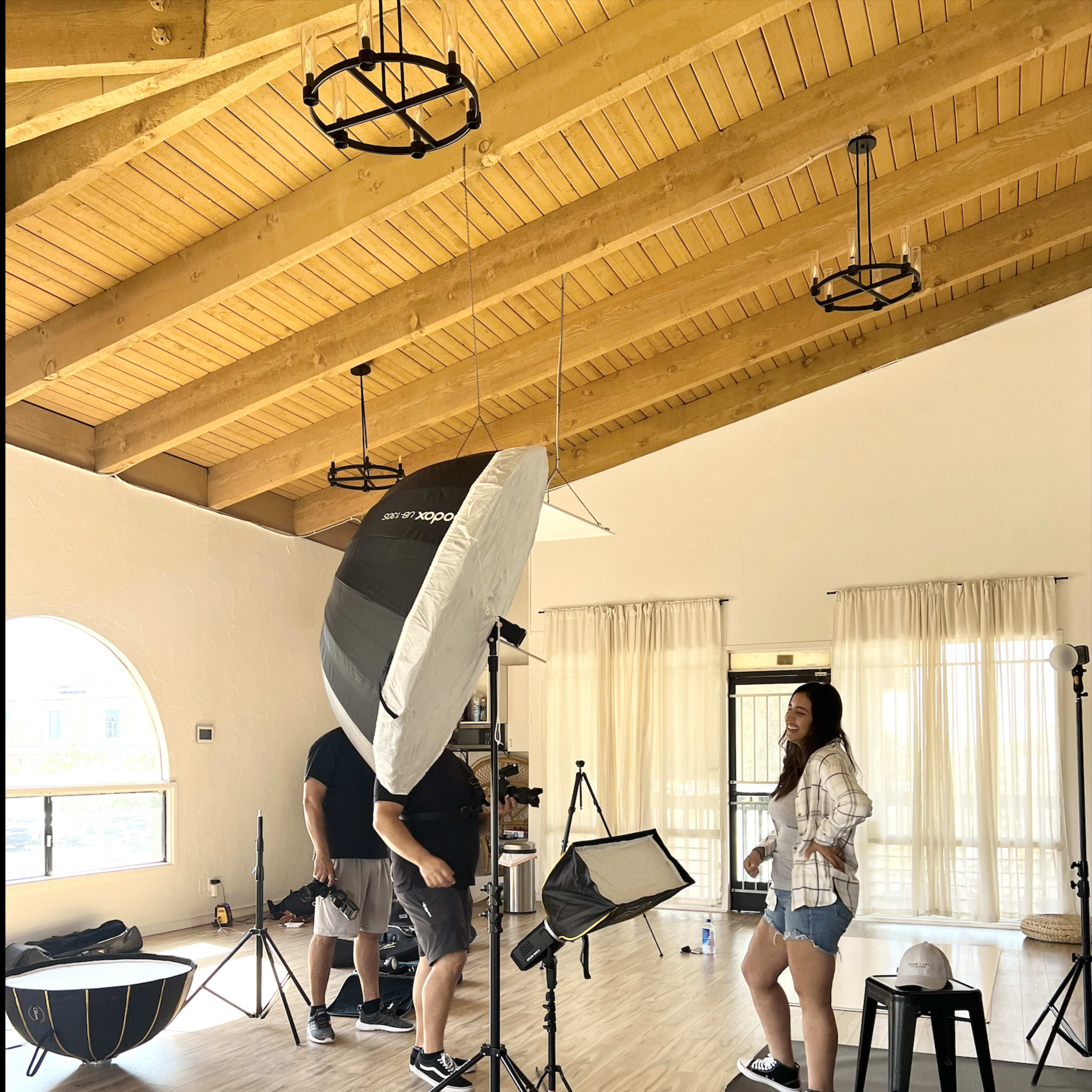 A photography studio features a model being photographed, with multiple lighting setups and equipment positioned around her.