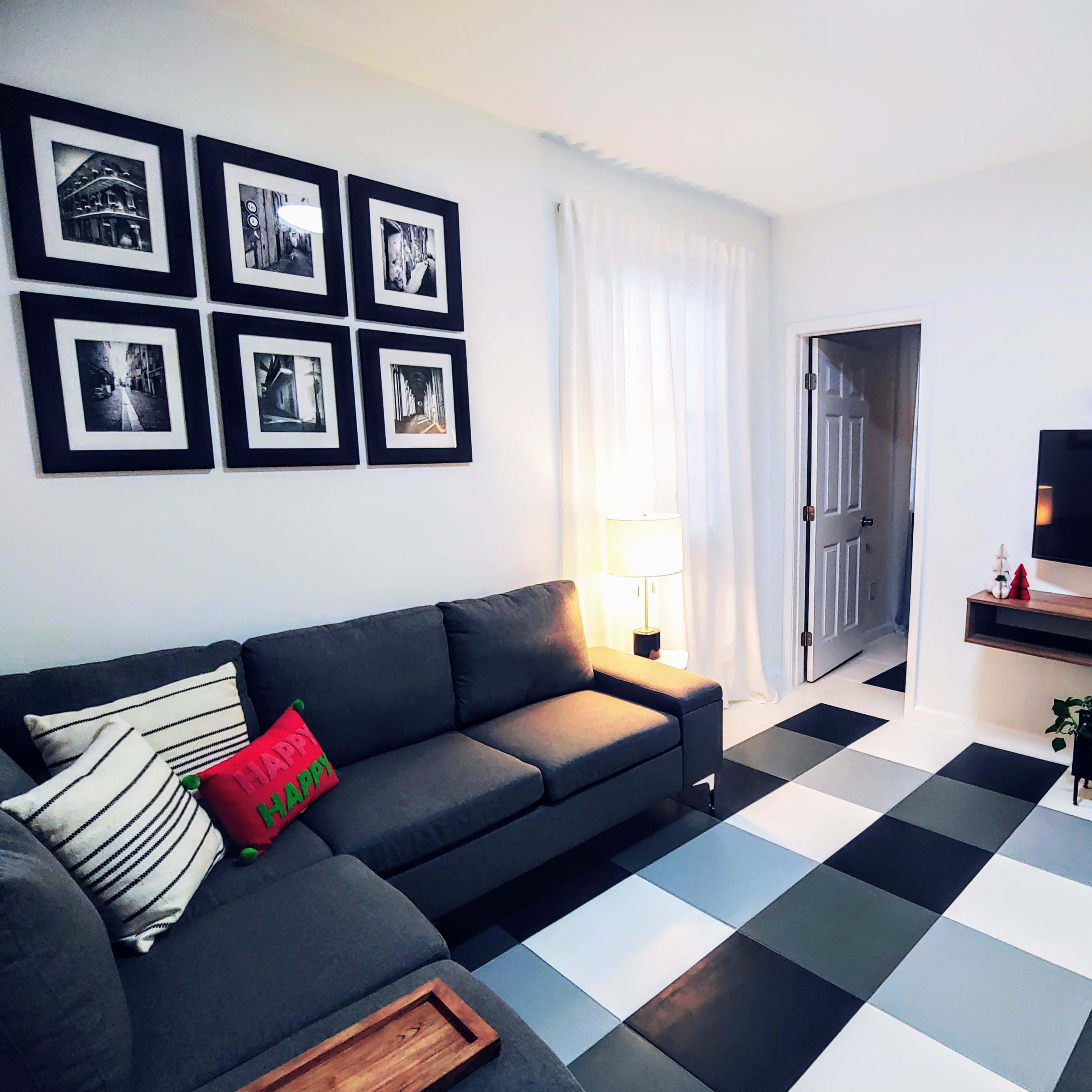 A modern living room features a gray sectional sofa, a wall with framed black and white photographs, a television mounted on a wooden stand, and a checkered floor in black, white, and gray.