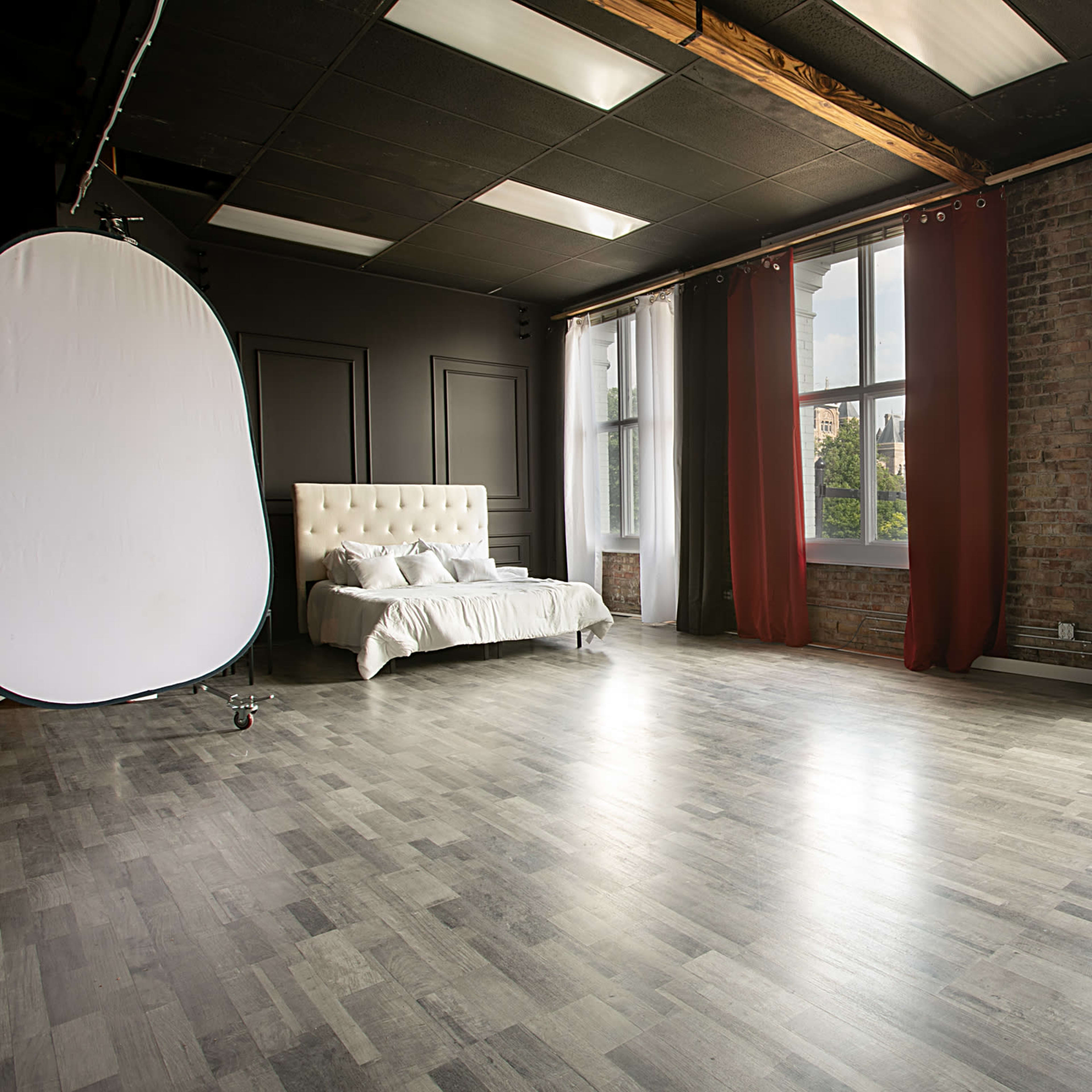 A spacious studio with a white bed, a tufted sofa, large windows with red and black curtains, and a light wooden floor.