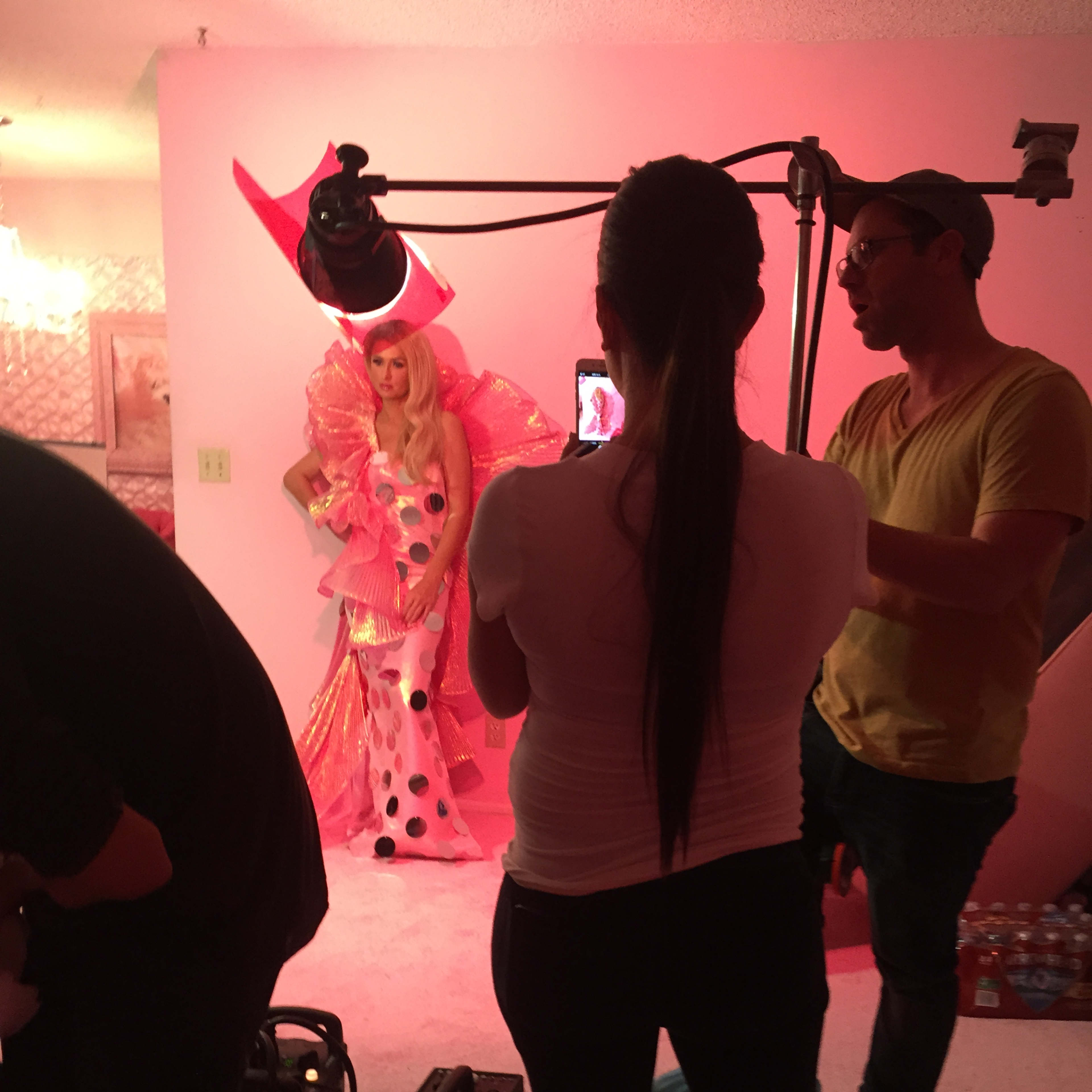 A model in a pink, polka-dotted costume poses against a pink backdrop while a crew member adjusts lighting and another holds a camera.