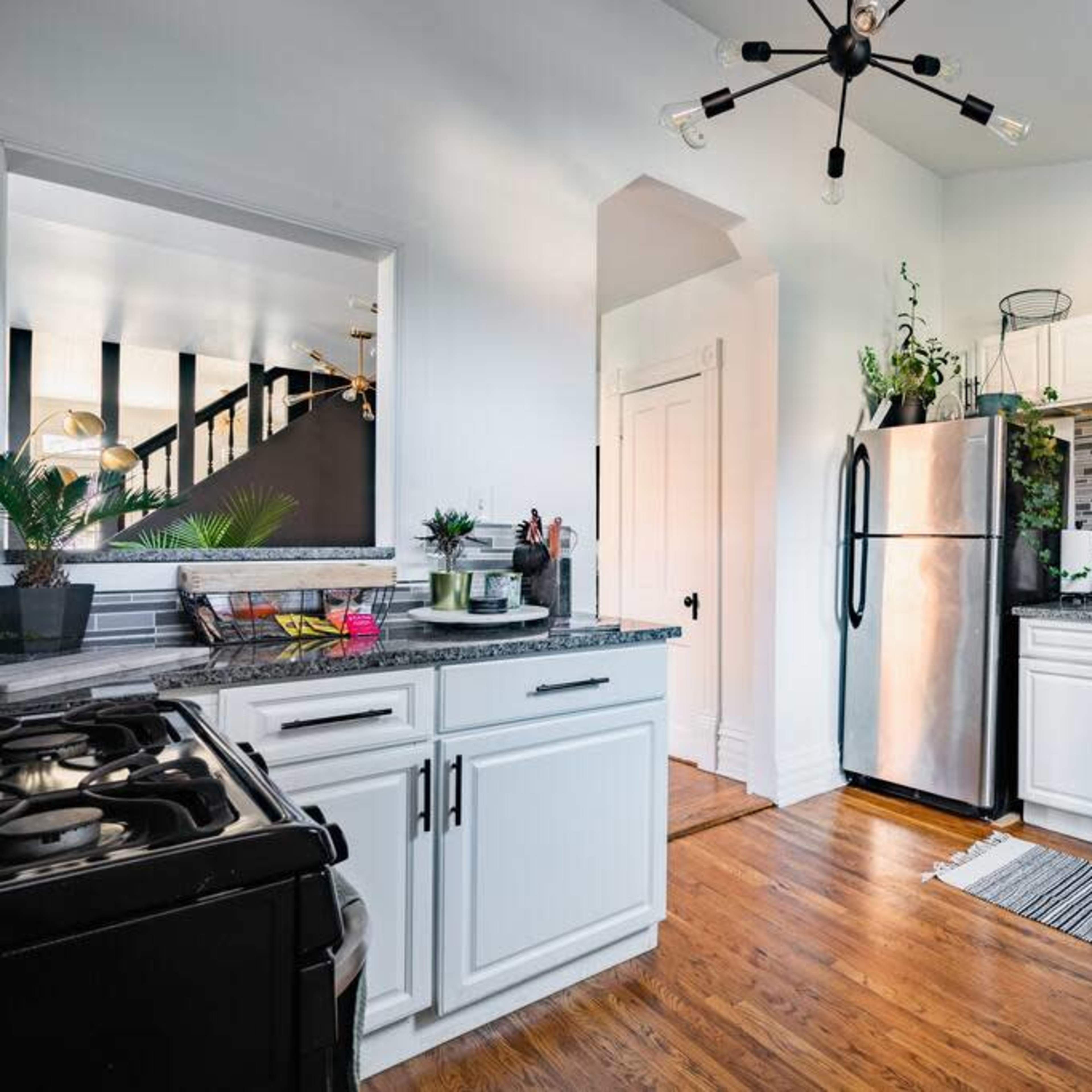 A modern kitchen with white cabinets, stainless steel appliances, and wooden floors, featuring a decorative backsplash and potted plants.