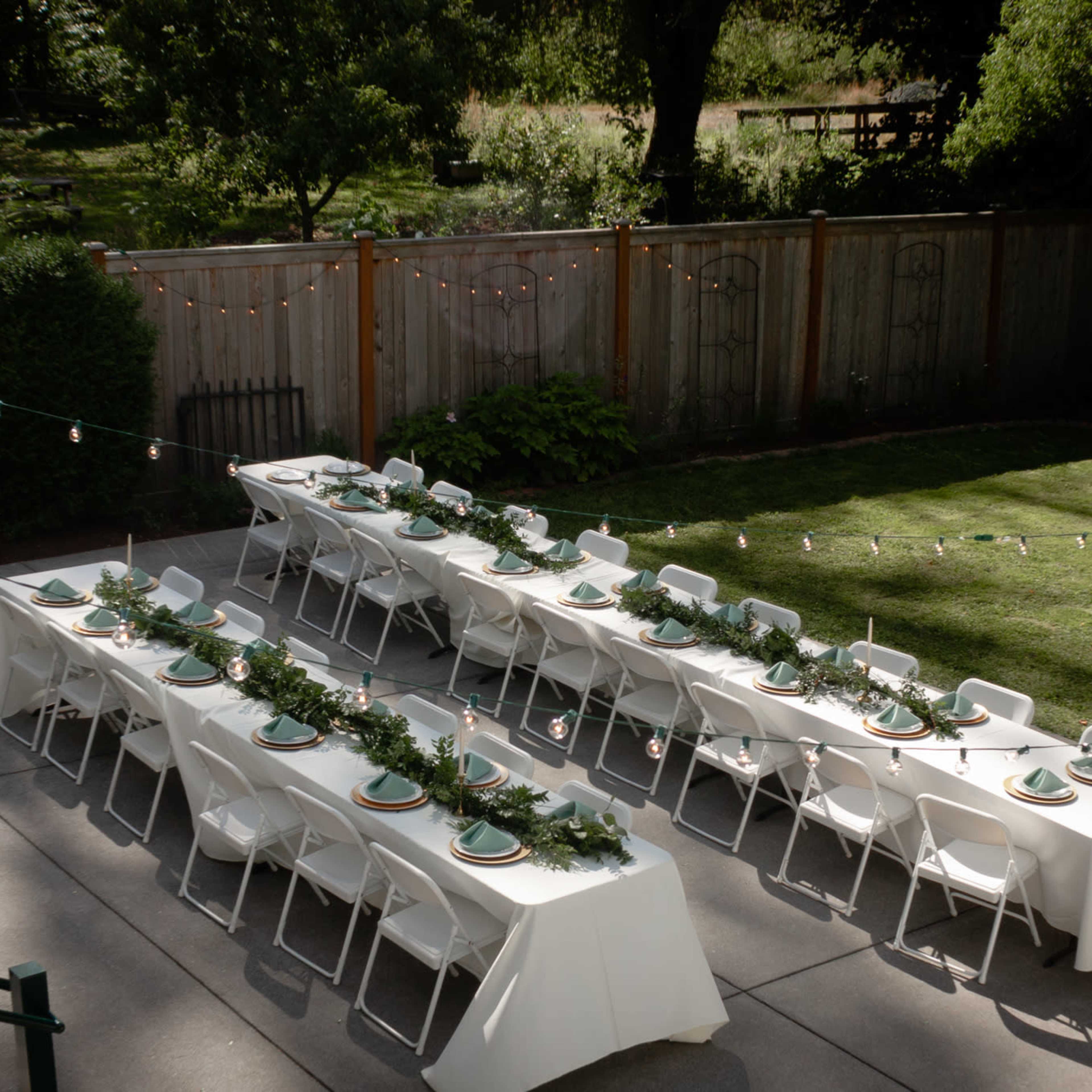 A garden setup with several long tables arranged in a row, each adorned with green table settings and surrounded by string lights.