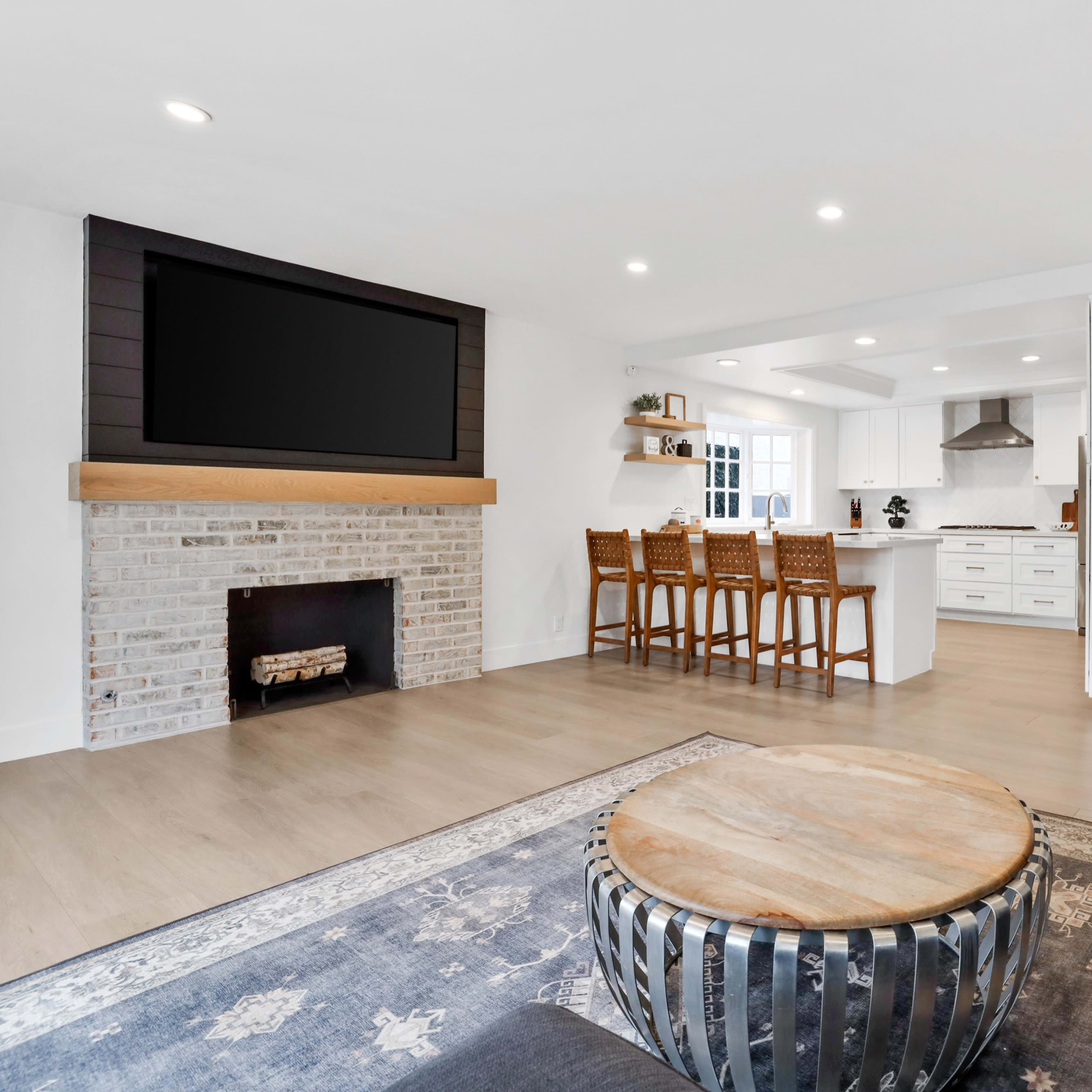 A modern living room features a brick fireplace, a large flat-screen TV mounted above a wooden mantel, and a circular coffee table surrounded by a dark sofa.